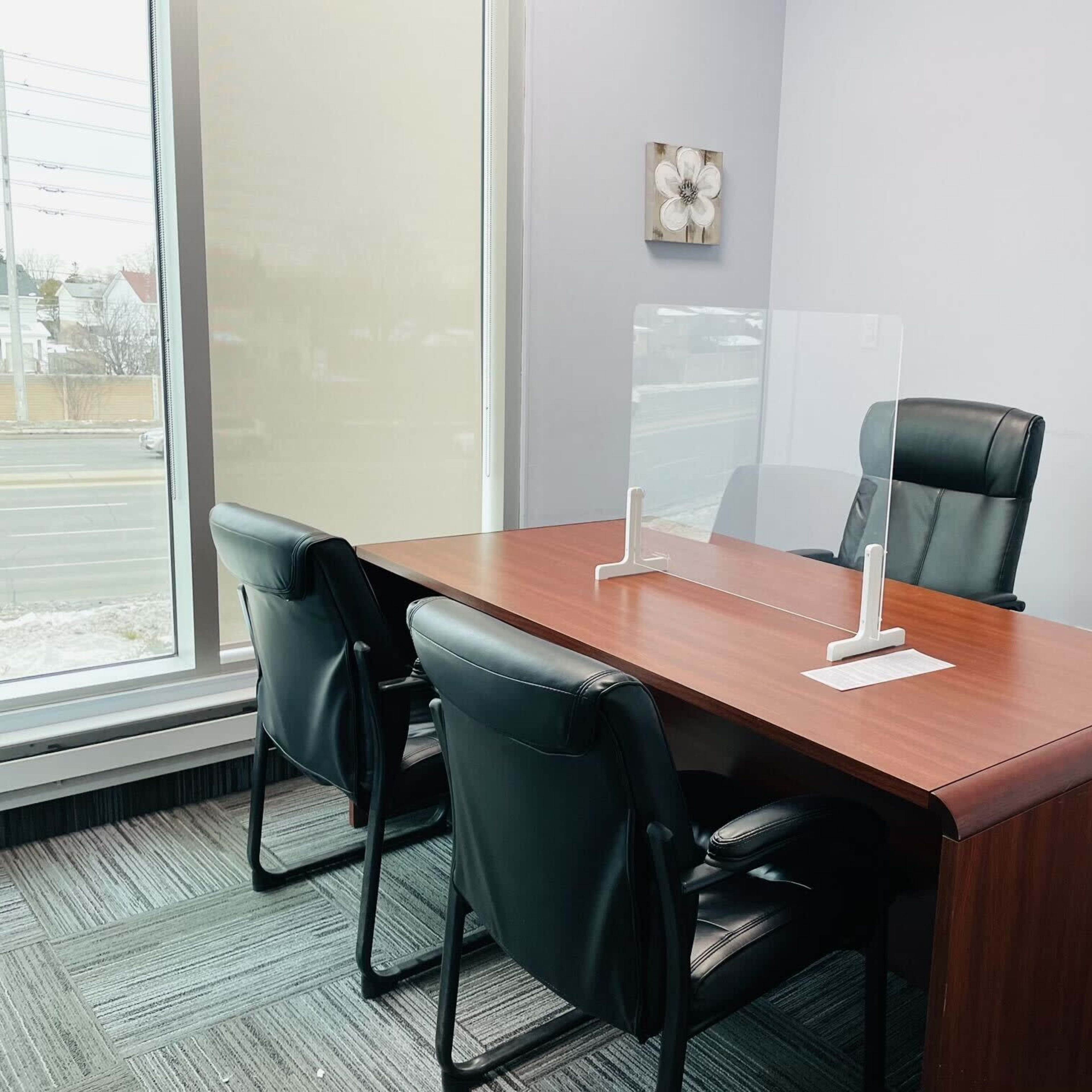 A modern office space features a wooden desk with a protective acrylic screen and two black chairs positioned around it.