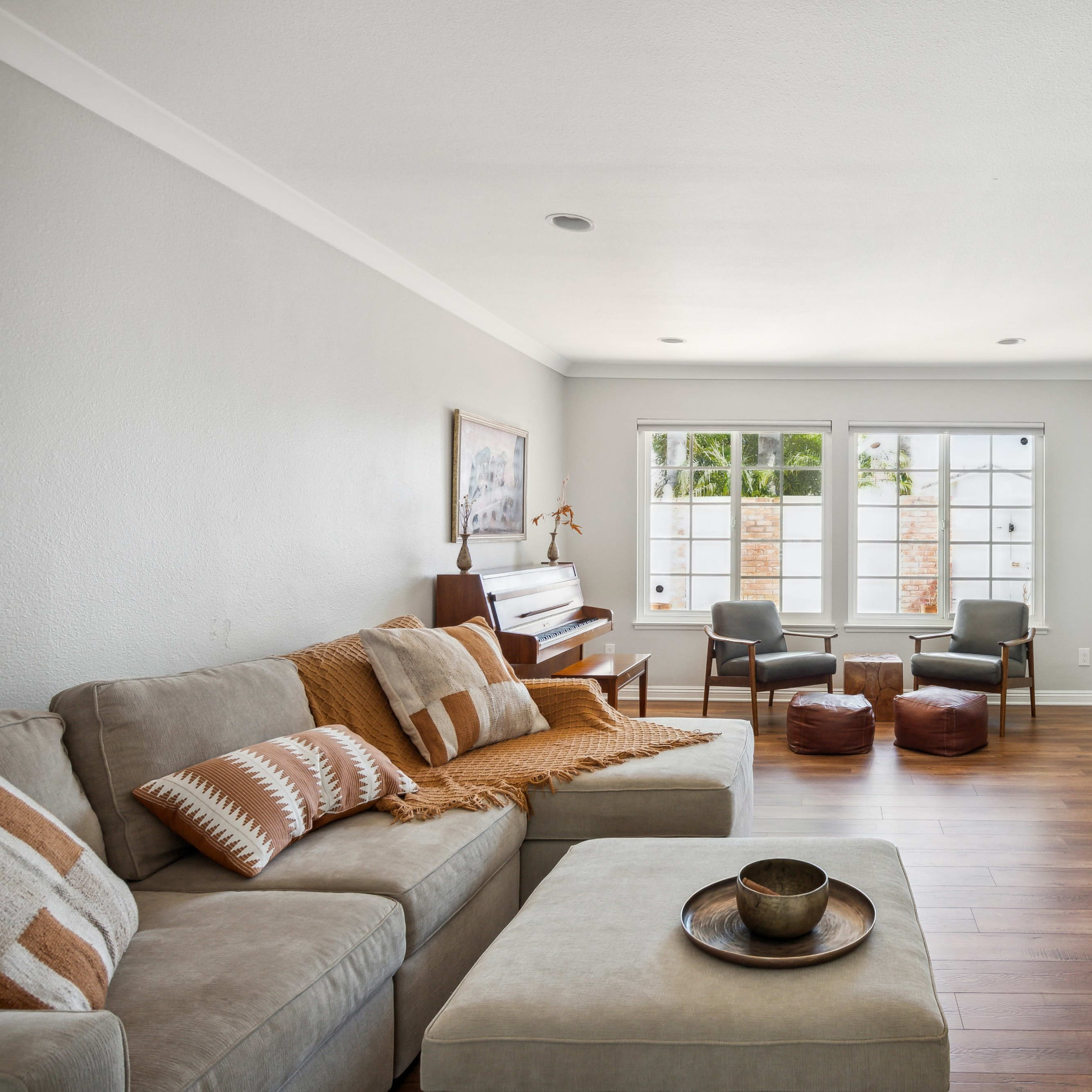 A light-filled living room features a sectional sofa, a piano, and two chairs with a coffee table in front of them.