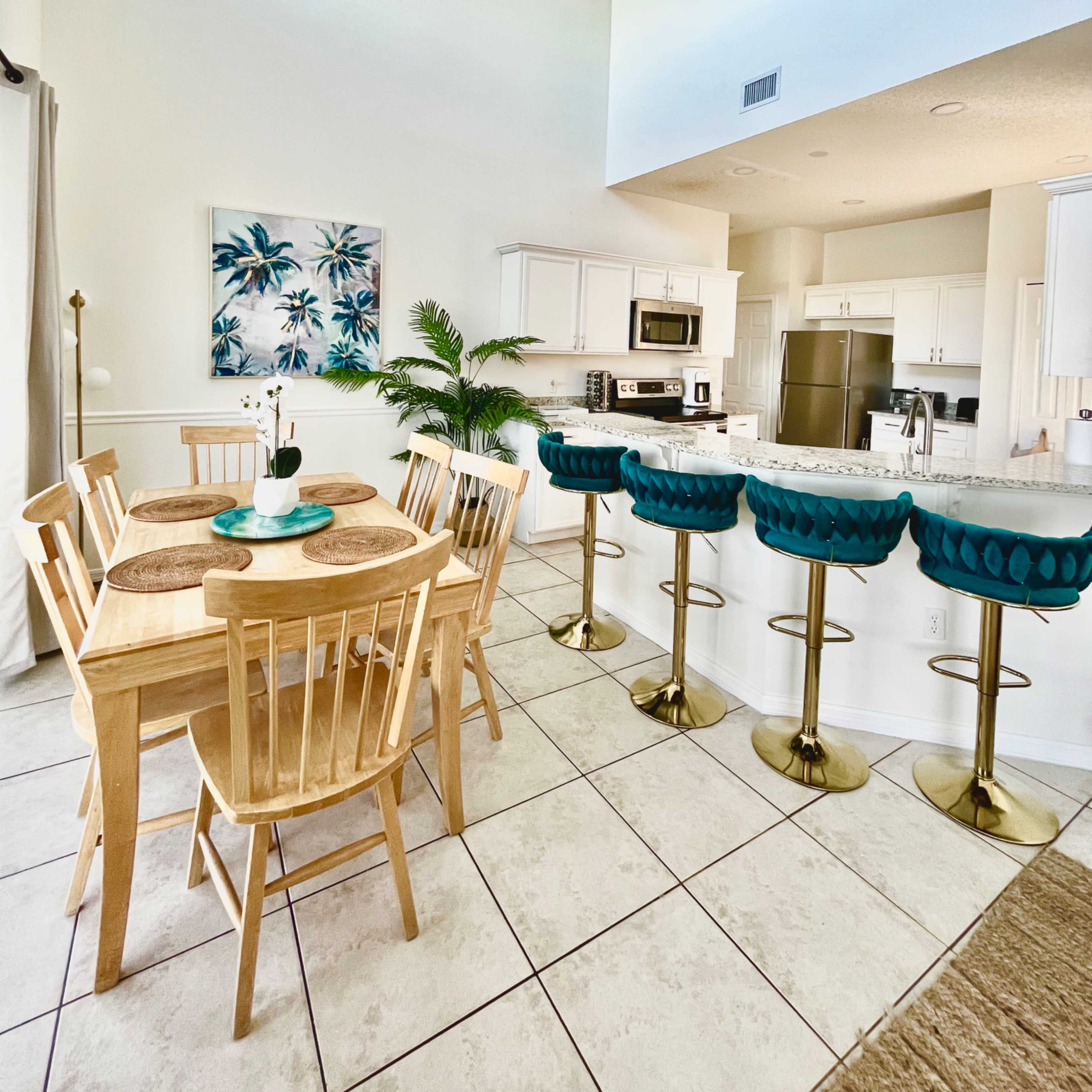 A modern kitchen and dining area feature a wooden table with four chairs, turquoise bar stools at a counter, and a decorative plant.