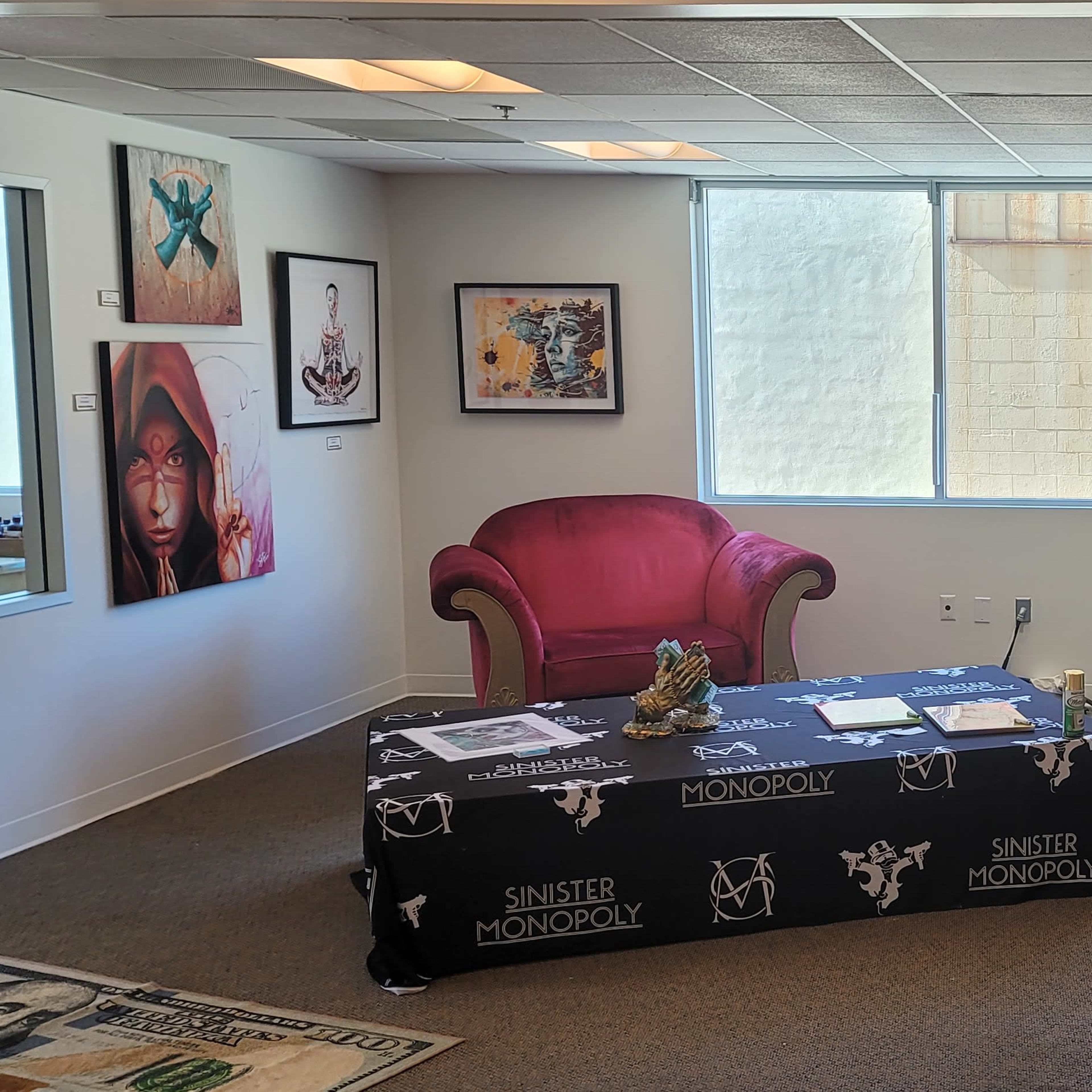 A modern lounge area with a pink sofa, a brown armchair, and a table covered in a black tablecloth featuring a Monopoly design, alongside wall art and a bar counter.