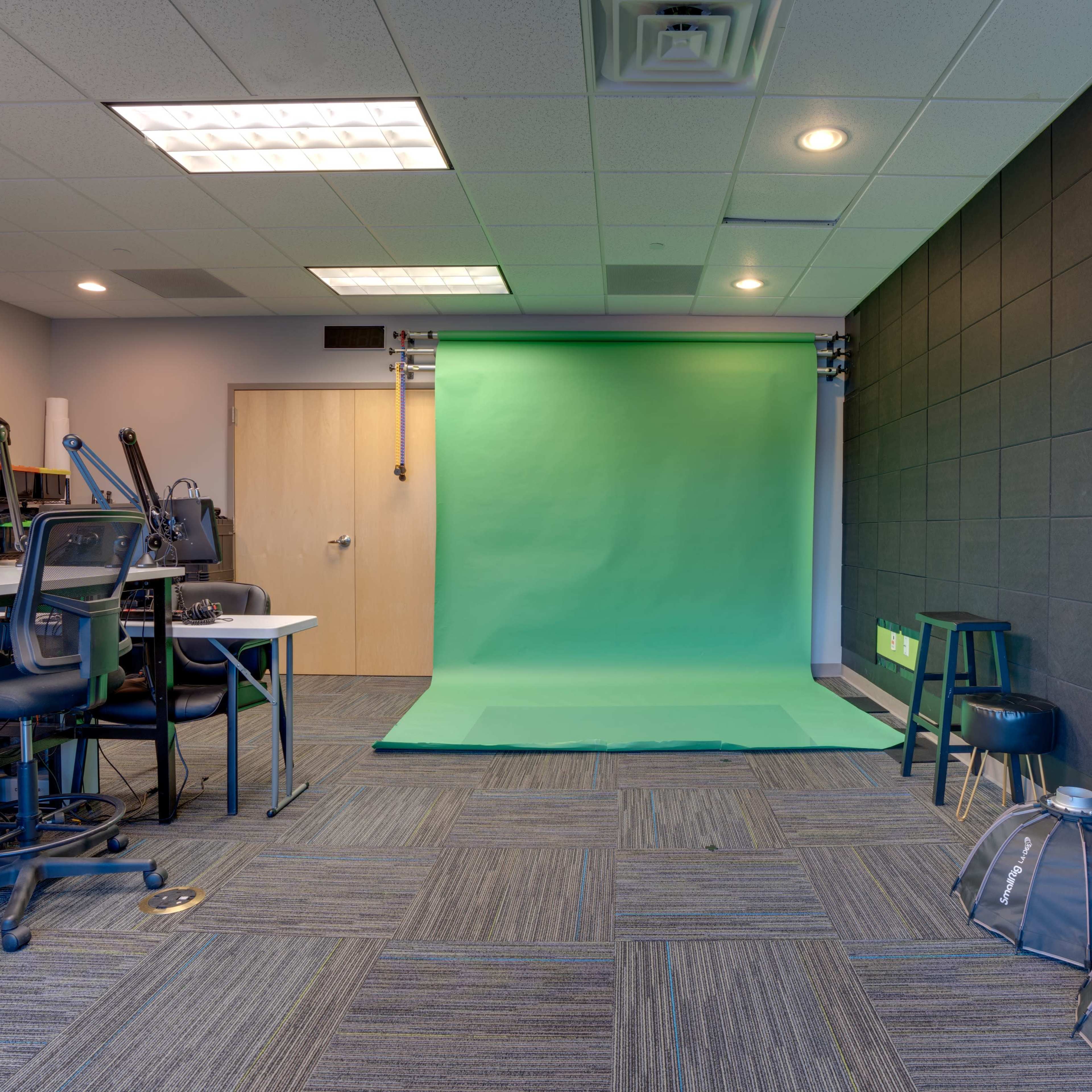 A room with a green backdrop and studio lights, accompanied by chairs and equipment on one side.