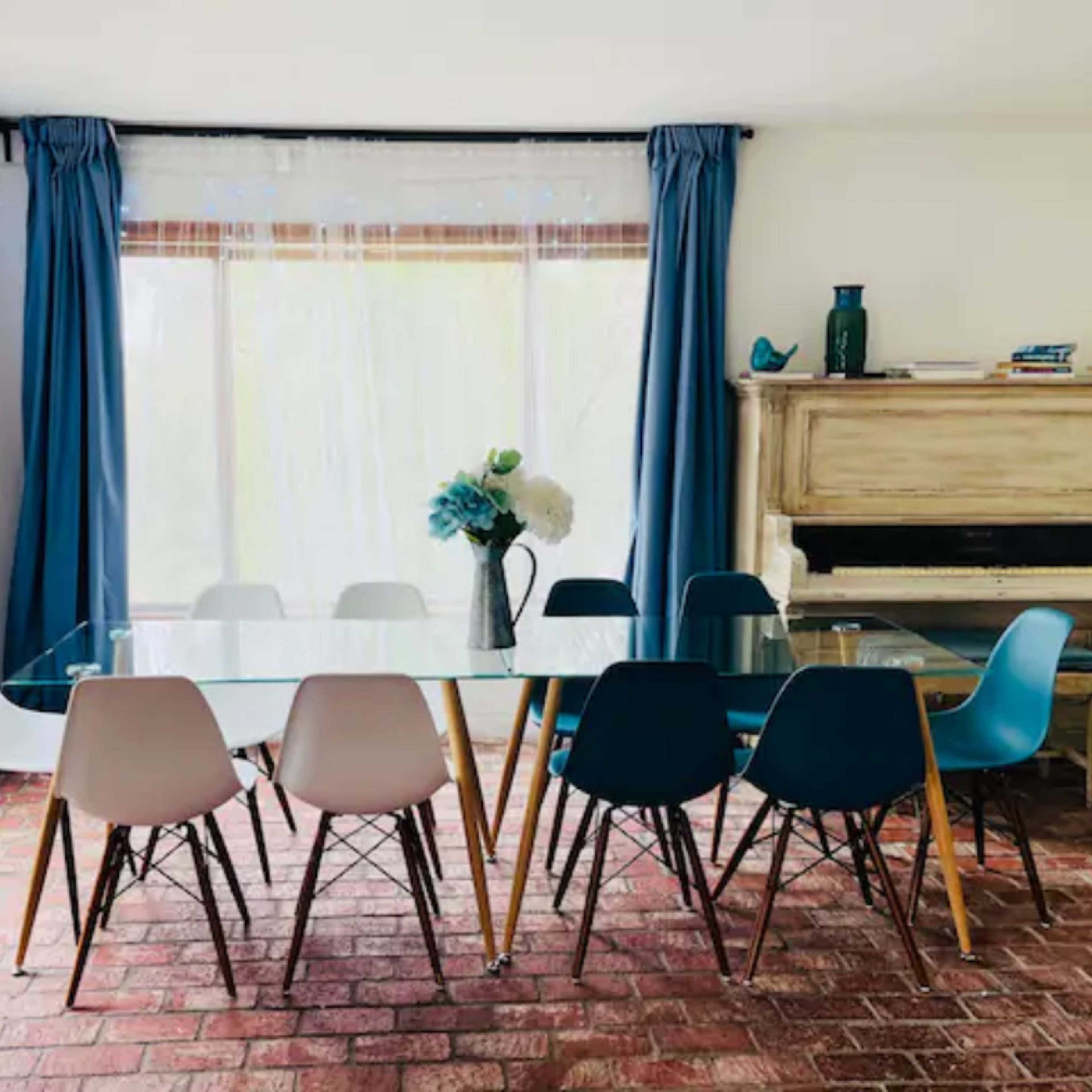 A bright dining room features a glass table surrounded by colorful chairs, with large windows draped in blue curtains and a piano in the corner.