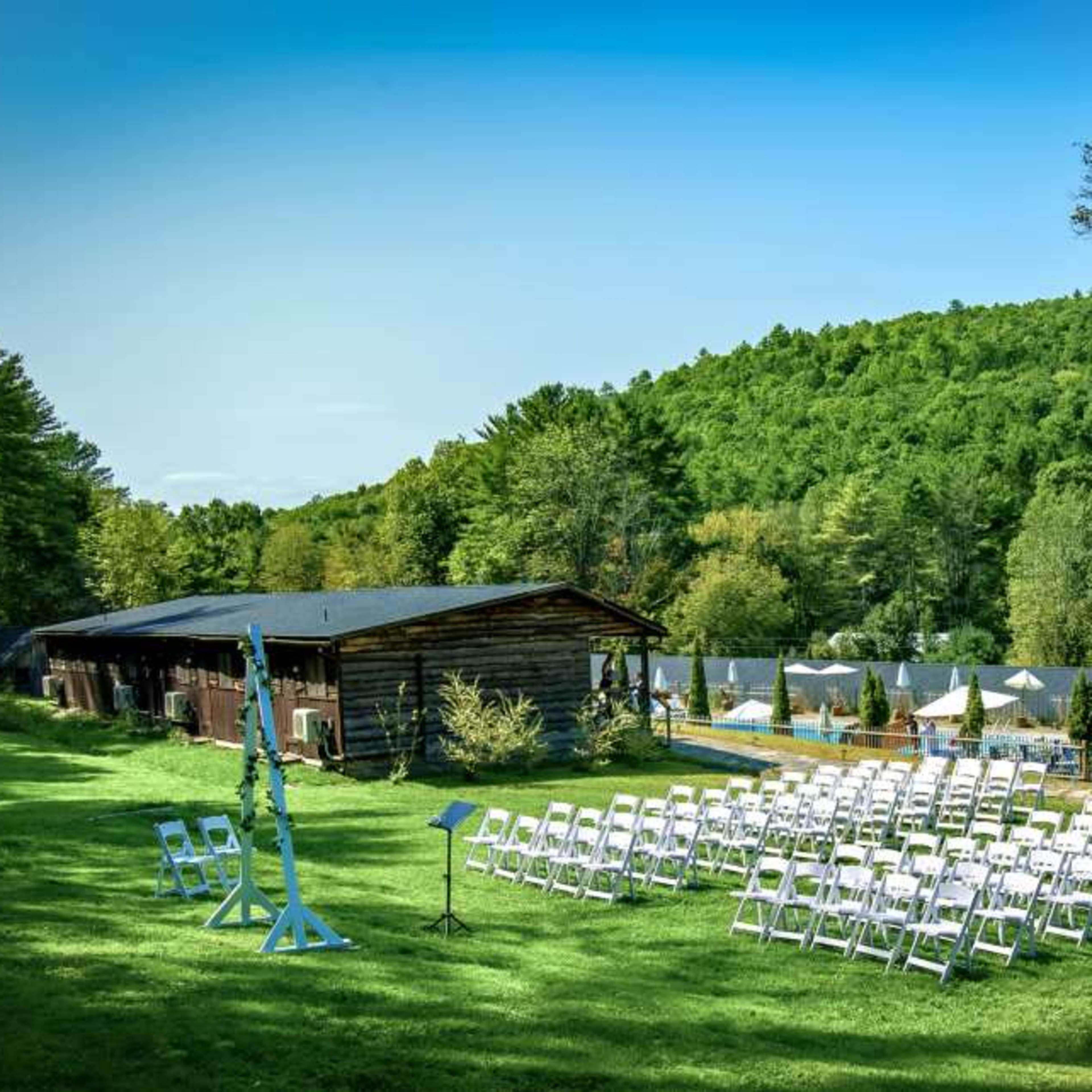A spacious outdoor venue is set up with rows of white chairs facing a grassy area, surrounded by trees and hills in the background.