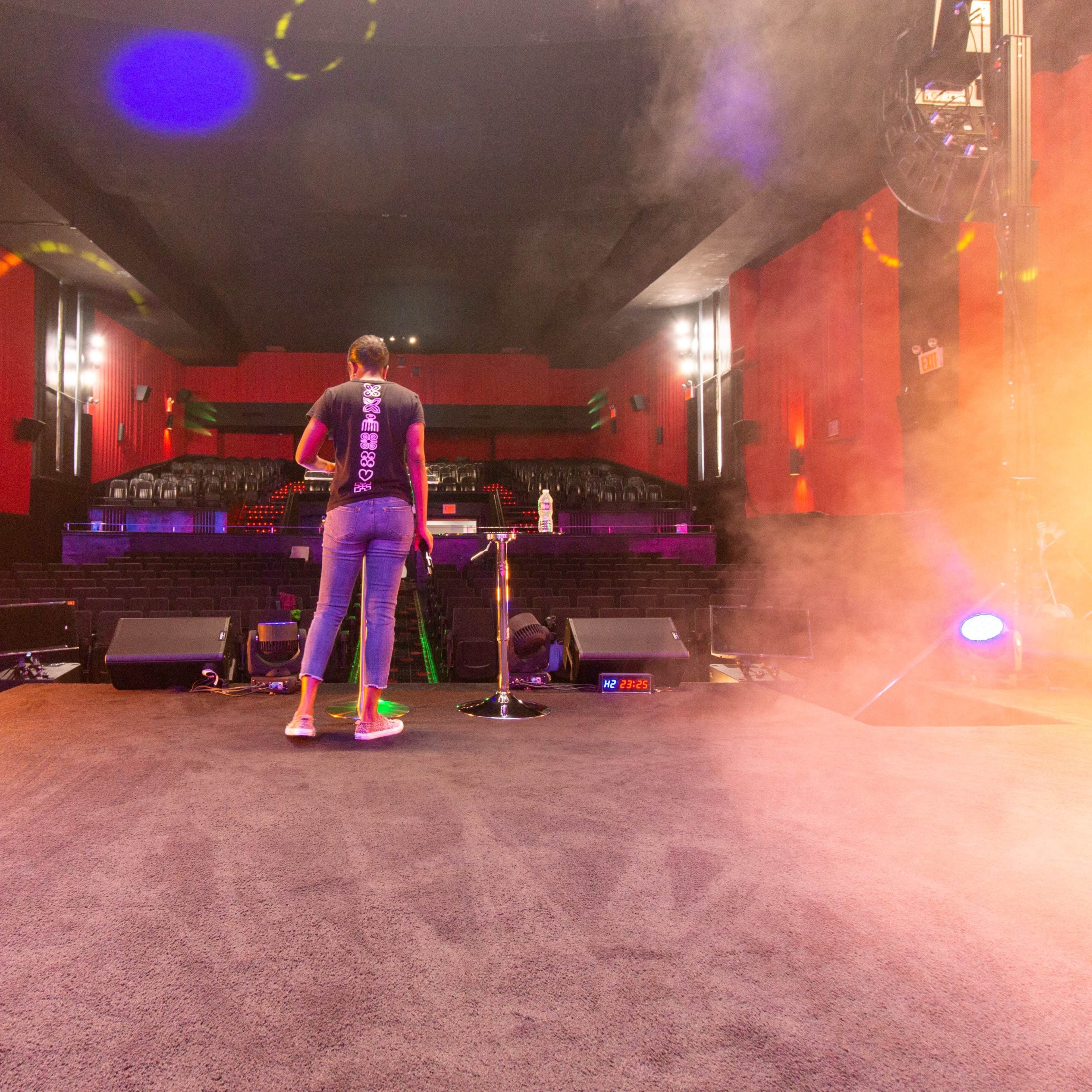 A performer stands on a stage in a theater with a microphone, facing an audience area illuminated by stage lights and surrounded by smoke.