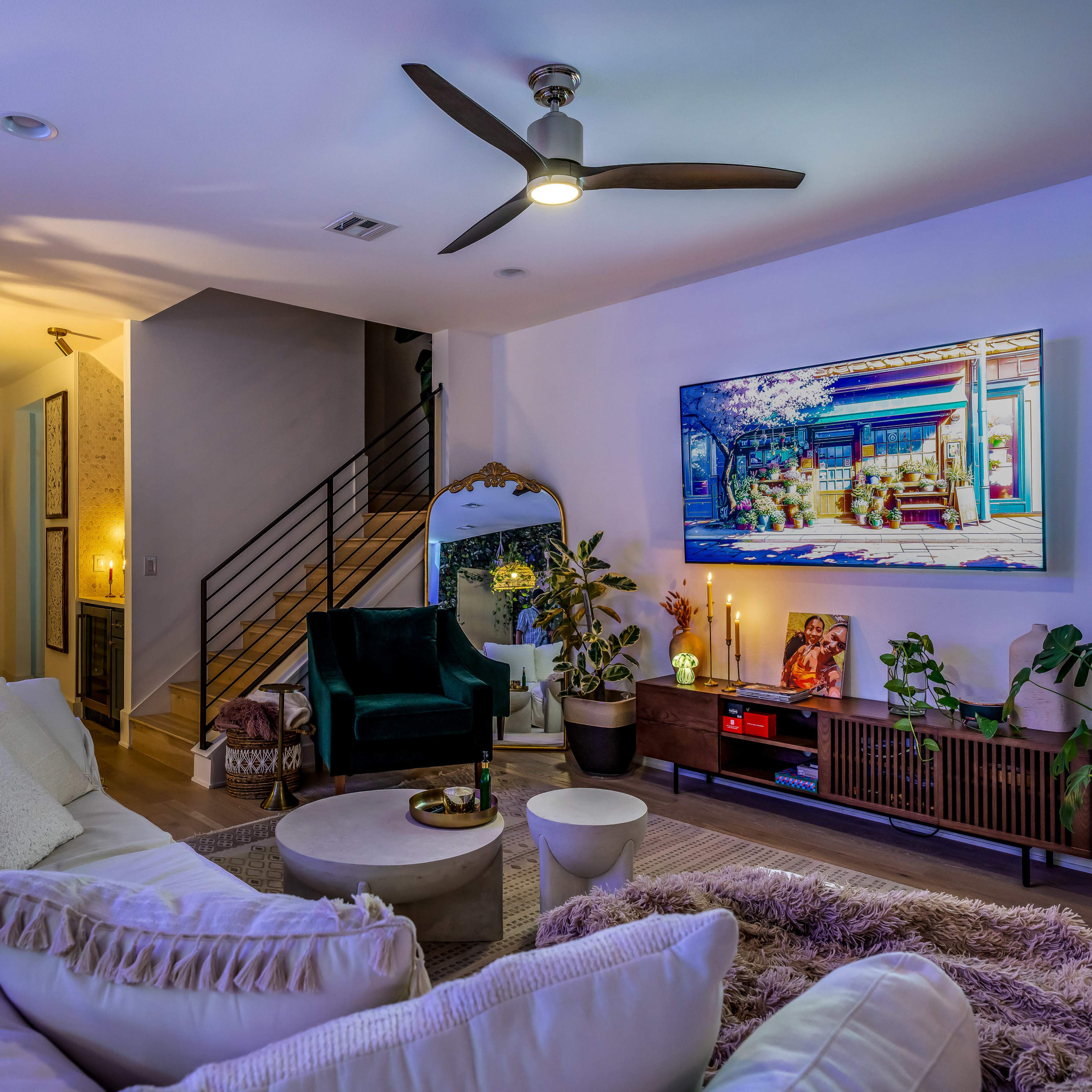 The living room features a white sectional sofa, a round coffee table, and a large TV mounted above a wooden sideboard, surrounded by indoor plants and soft lighting.