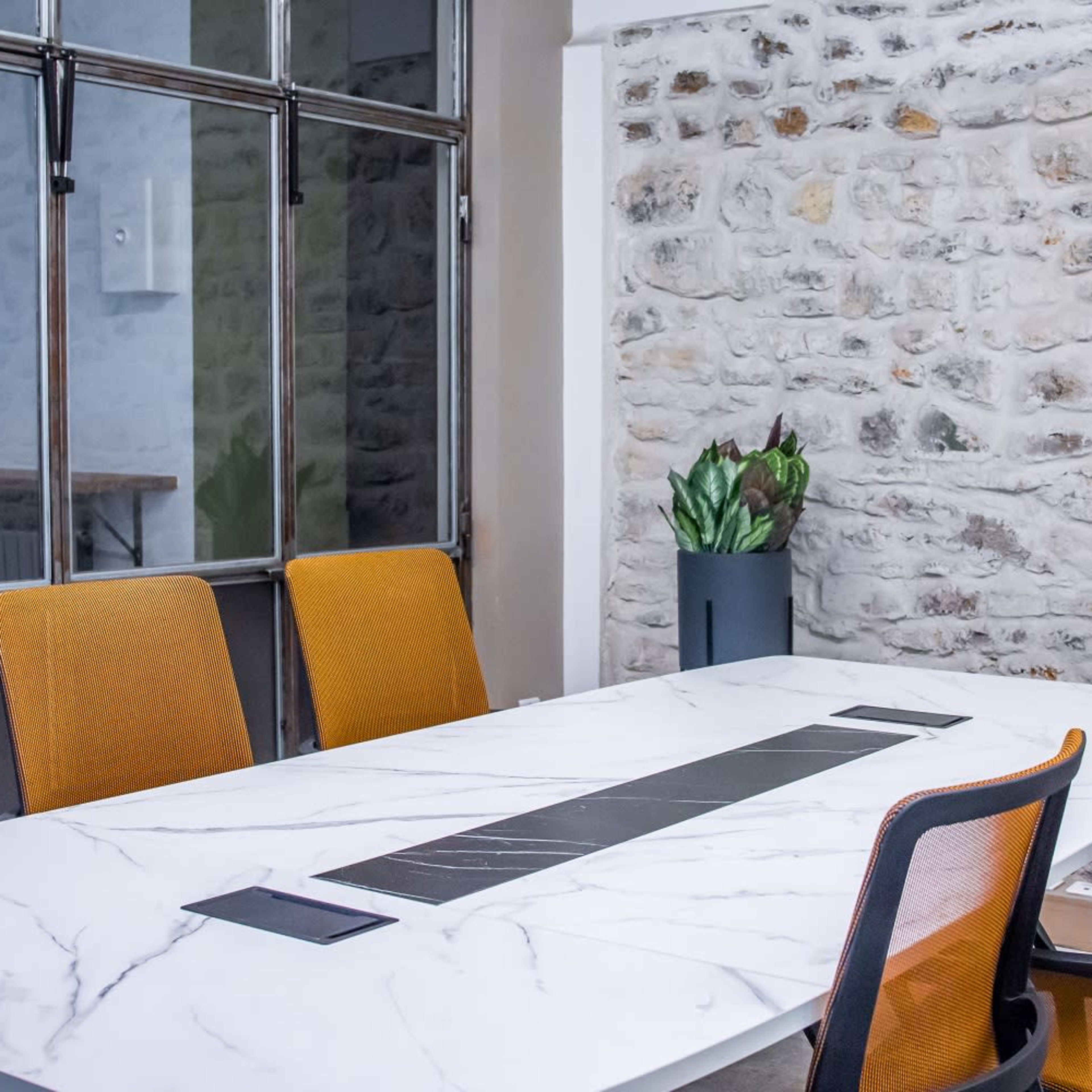 A conference room features a marble table with orange chairs, set against a stone wall and large windows.