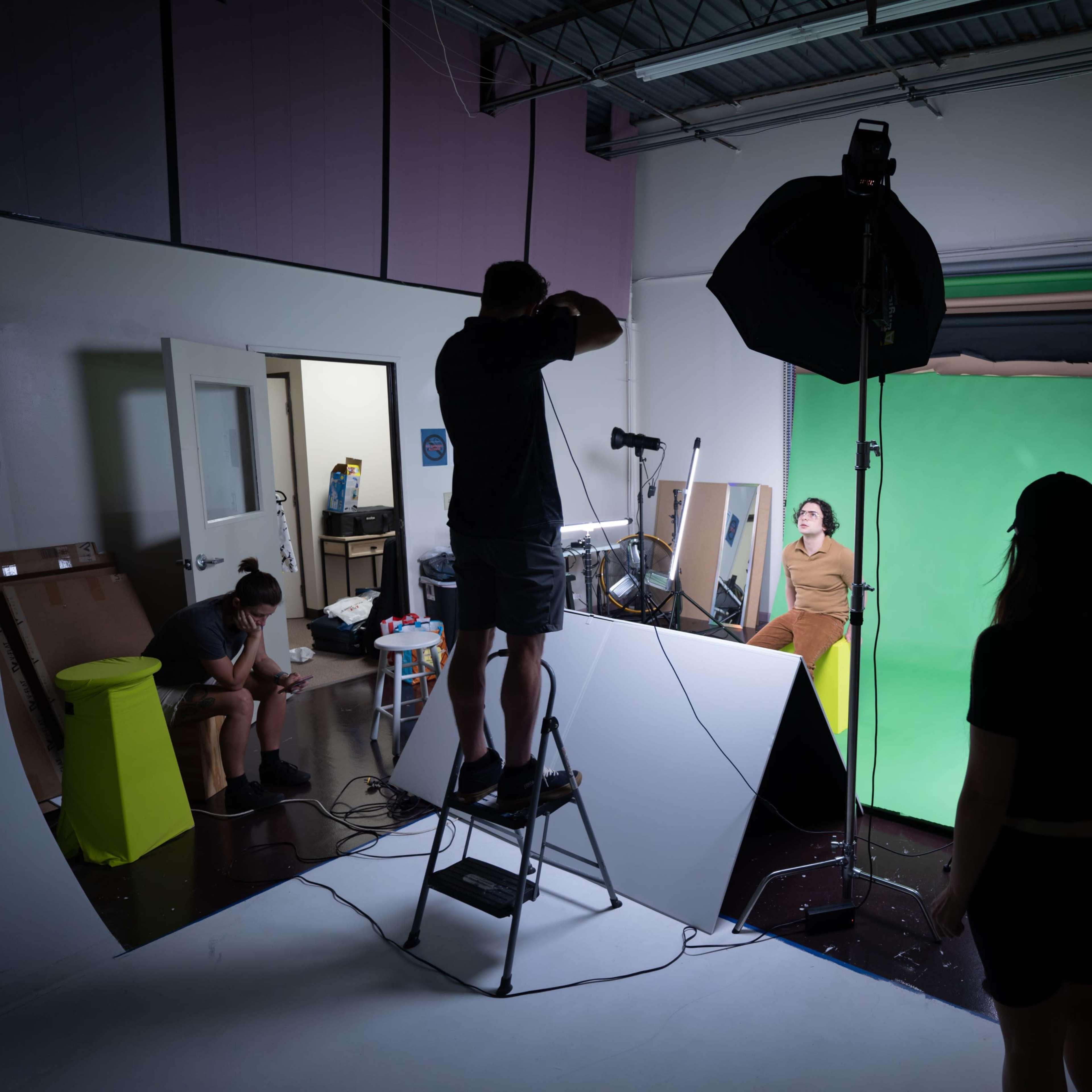 A photography studio is set up with a model posing on a set, while photographers and assistants adjust lighting and equipment around them.