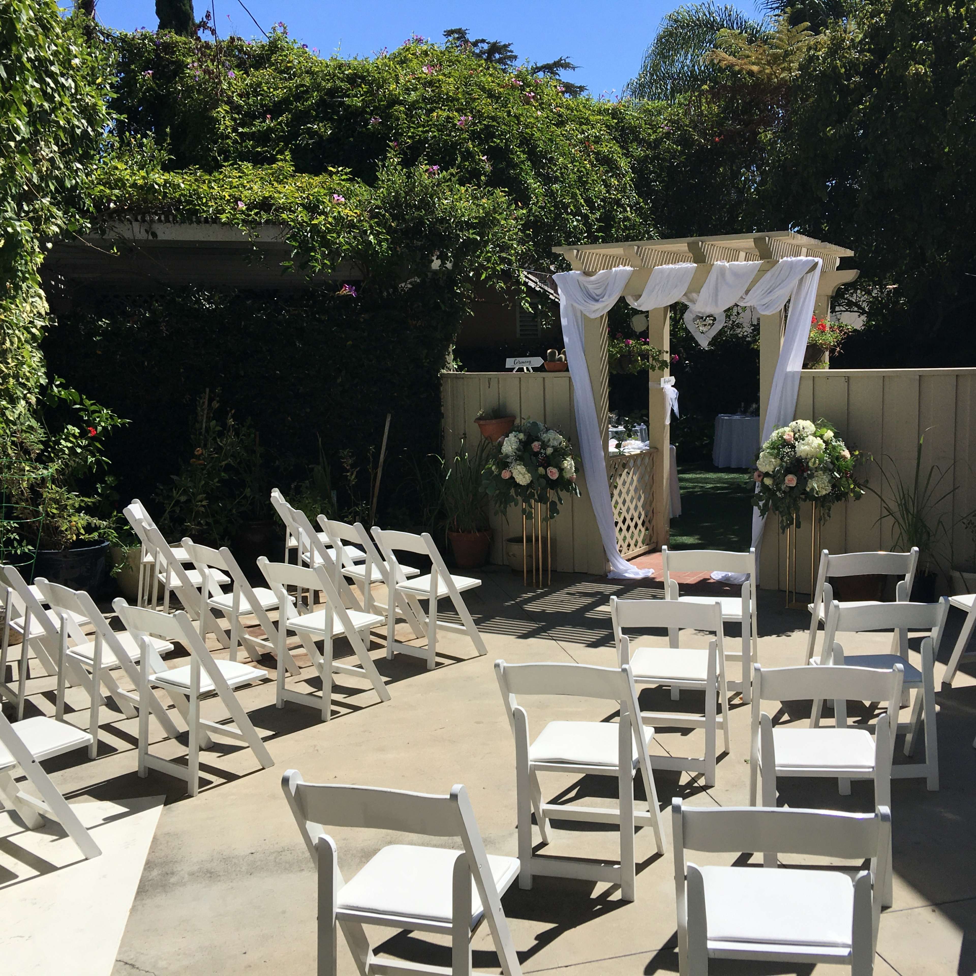 A small outdoor wedding setup with white chairs arranged in rows facing a decorated arch surrounded by flowers.