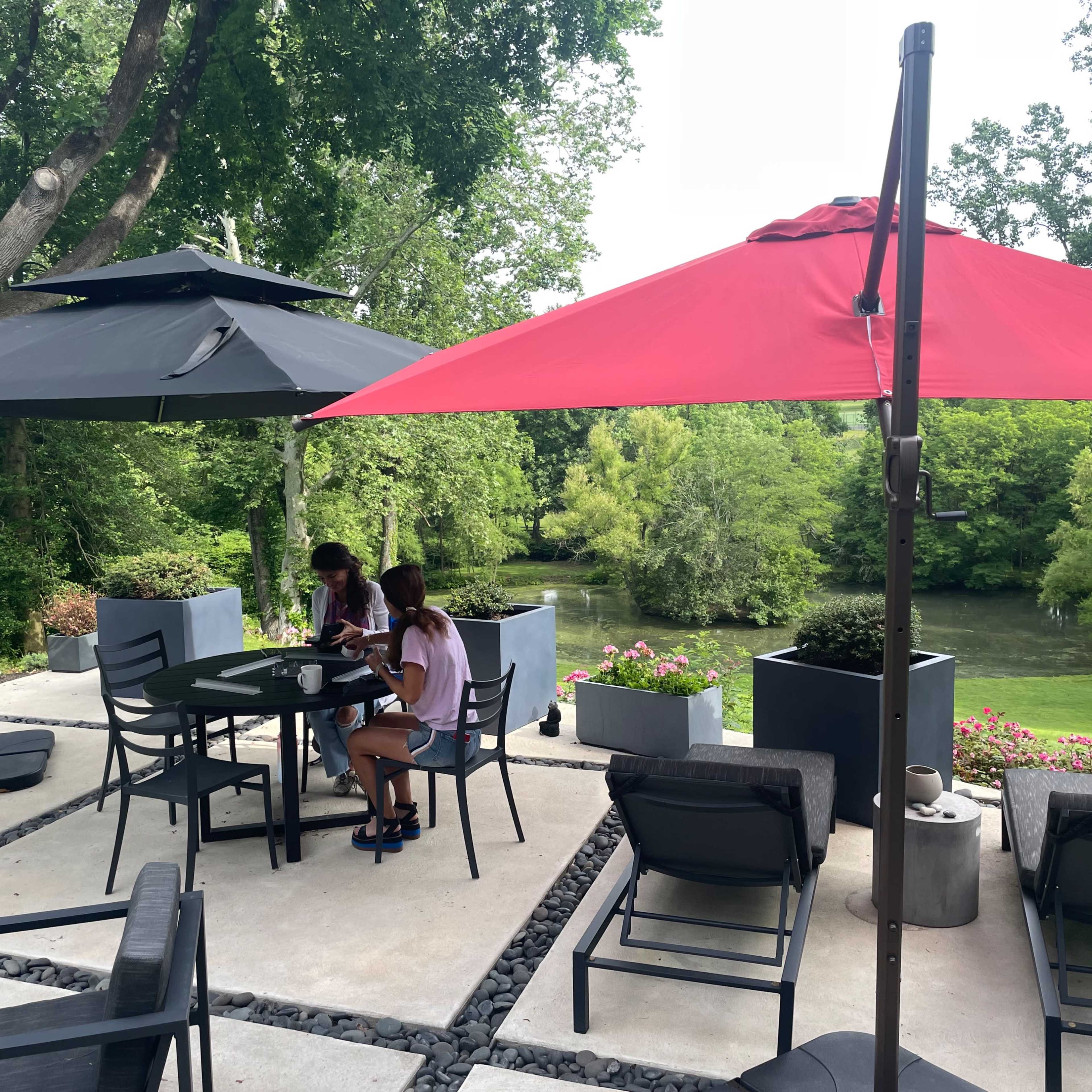 Two people sit at a table under a black and a red umbrella beside a river, with greenery in the background.