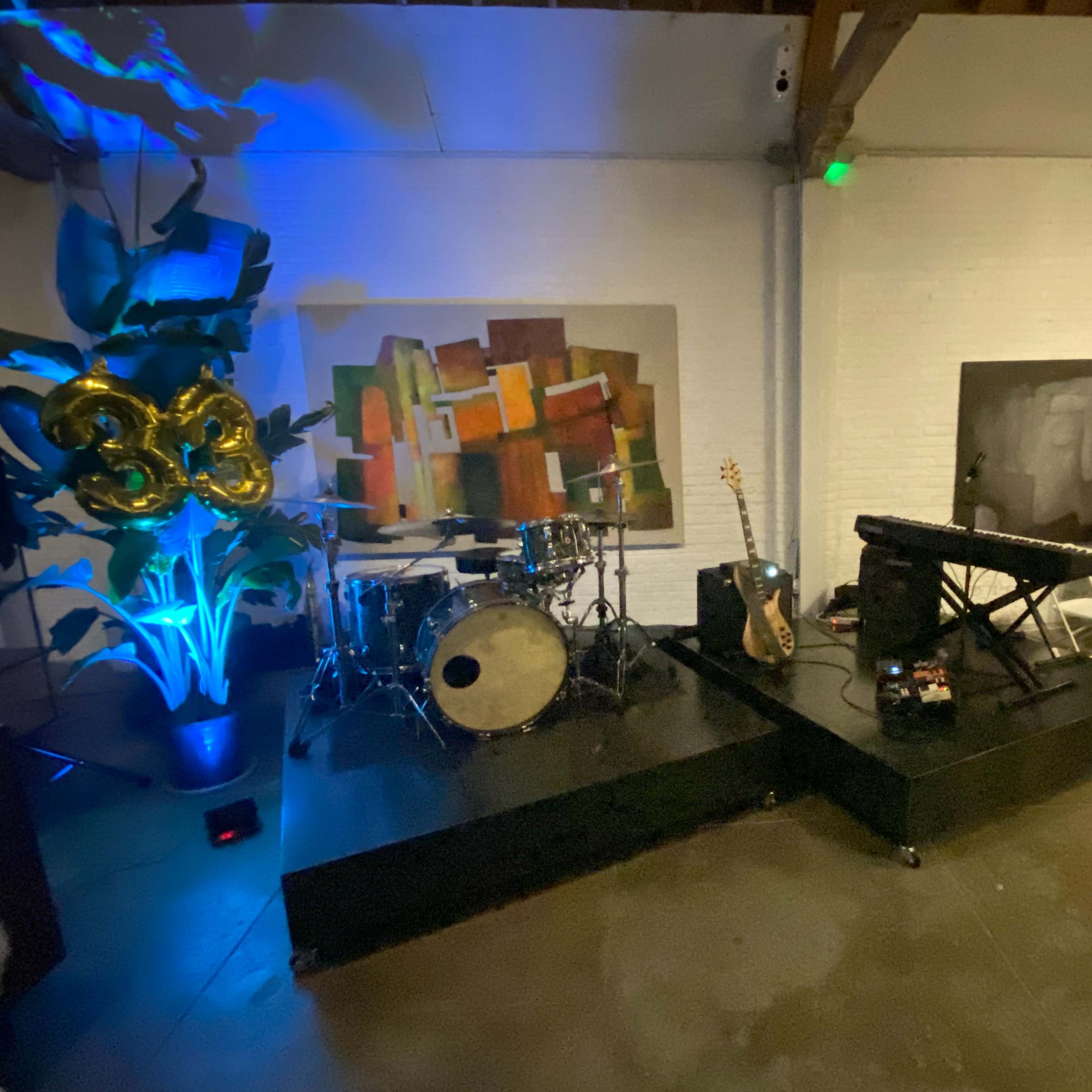 A small stage setup with musical instruments, including a drum set and keyboard, alongside decorative balloons and seating for guests in a dimly lit venue.