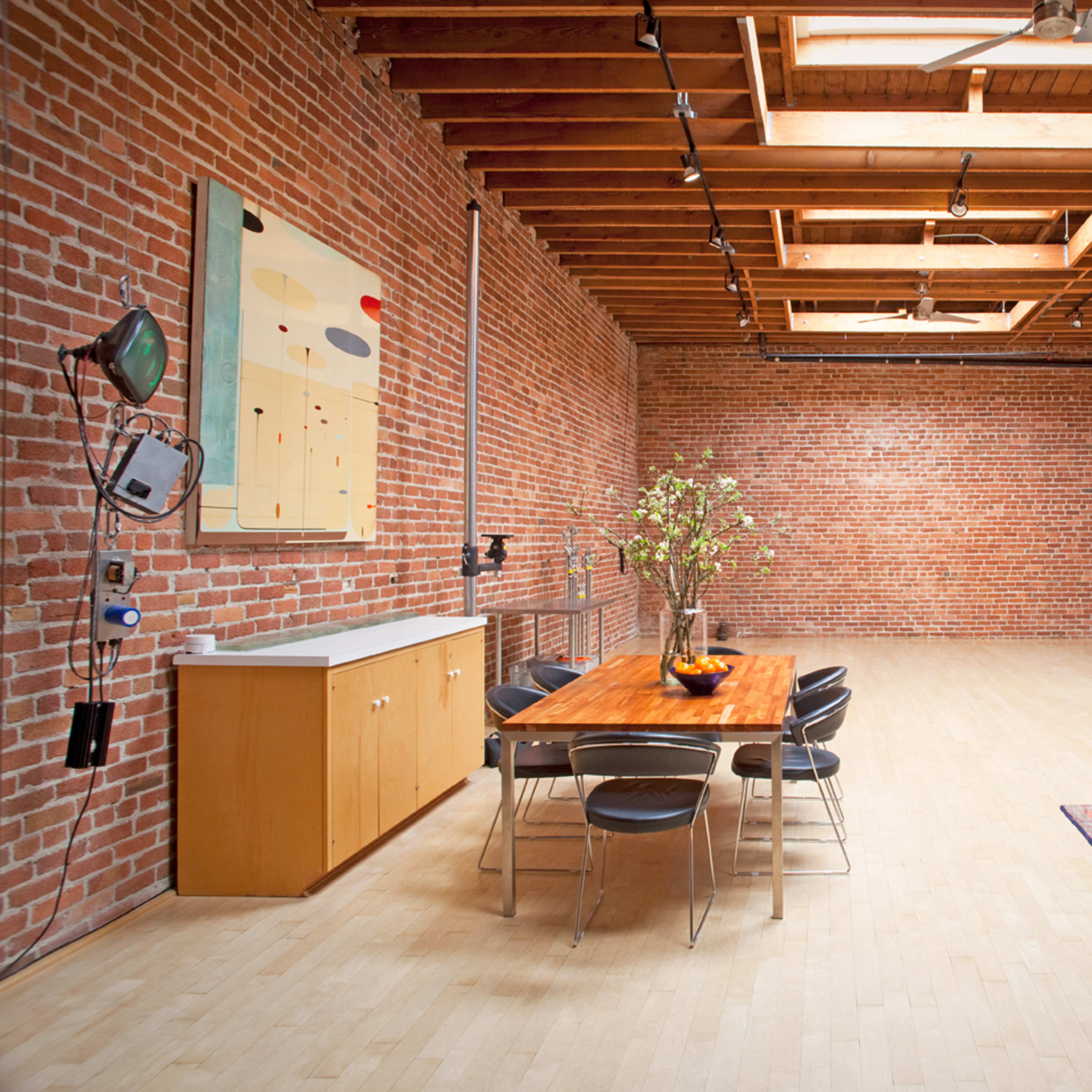 A spacious, industrial-style room with exposed brick walls, overhead beams, a wooden dining table surrounded by black chairs, a leather sofa, and an artwork display.