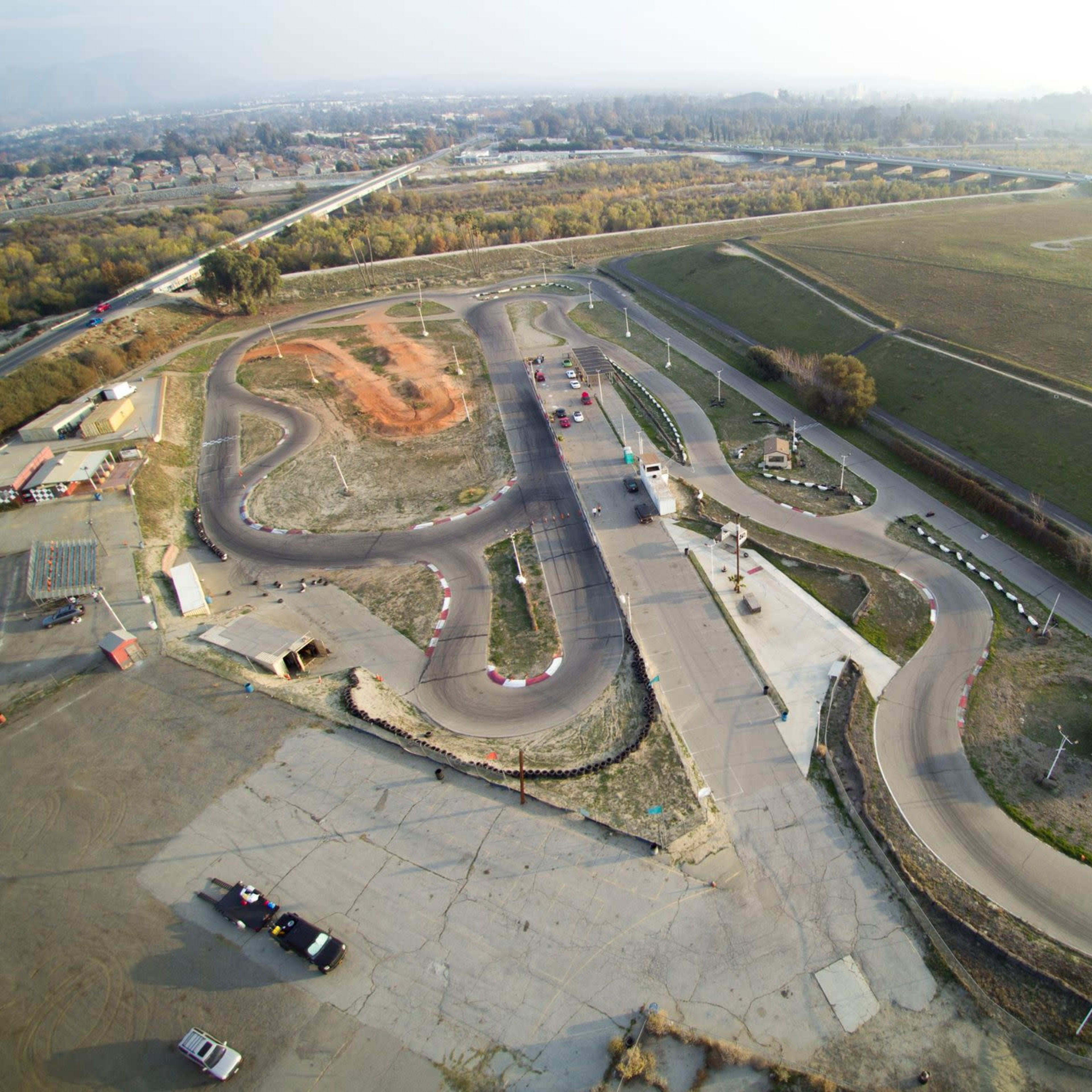 An aerial view of a kart racing track with several curves and a partially dirt area, surrounded by grassy fields and residential areas.