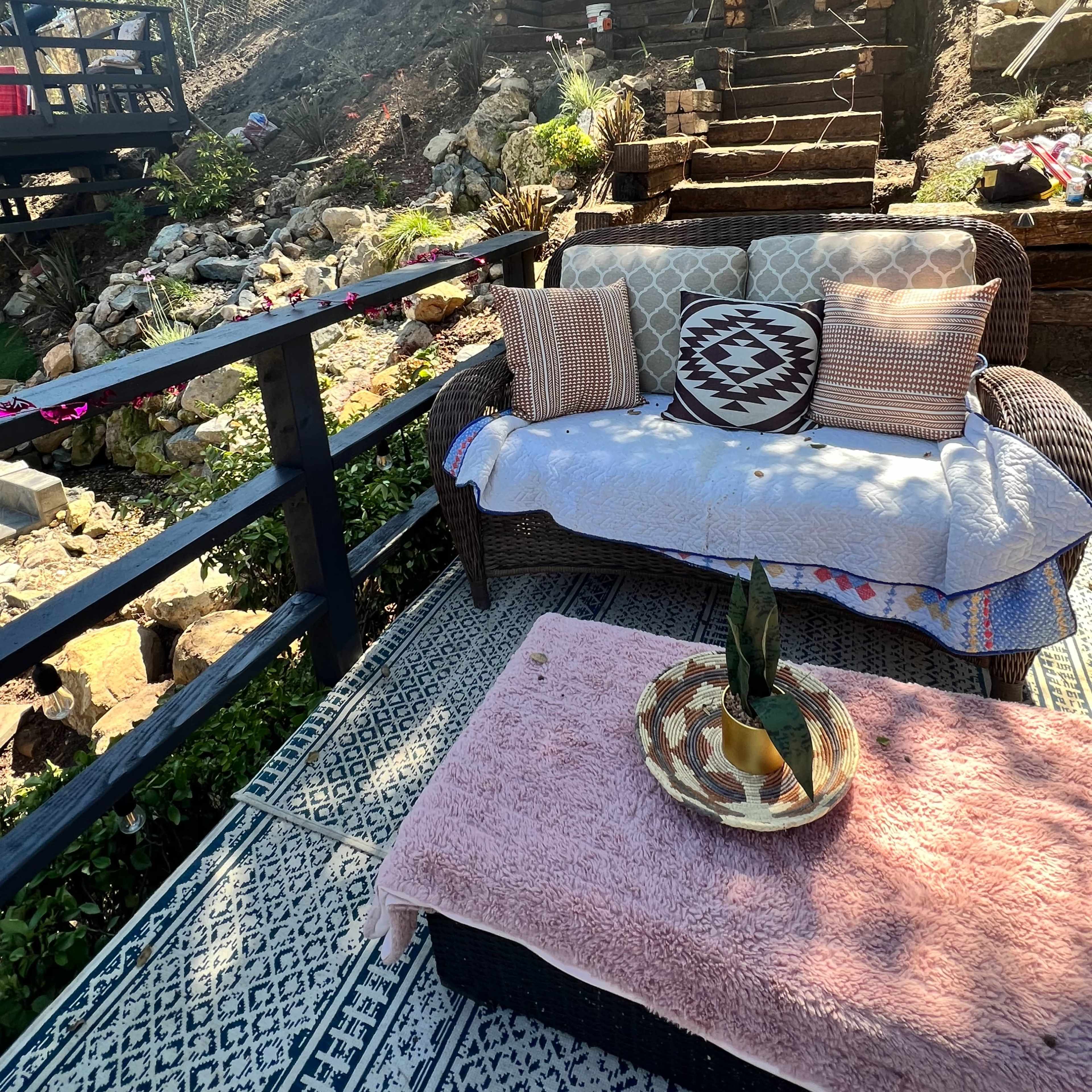 A cozy seating area with a sofa and a decorative table, surrounded by a hillside garden and a wooden deck in the background.