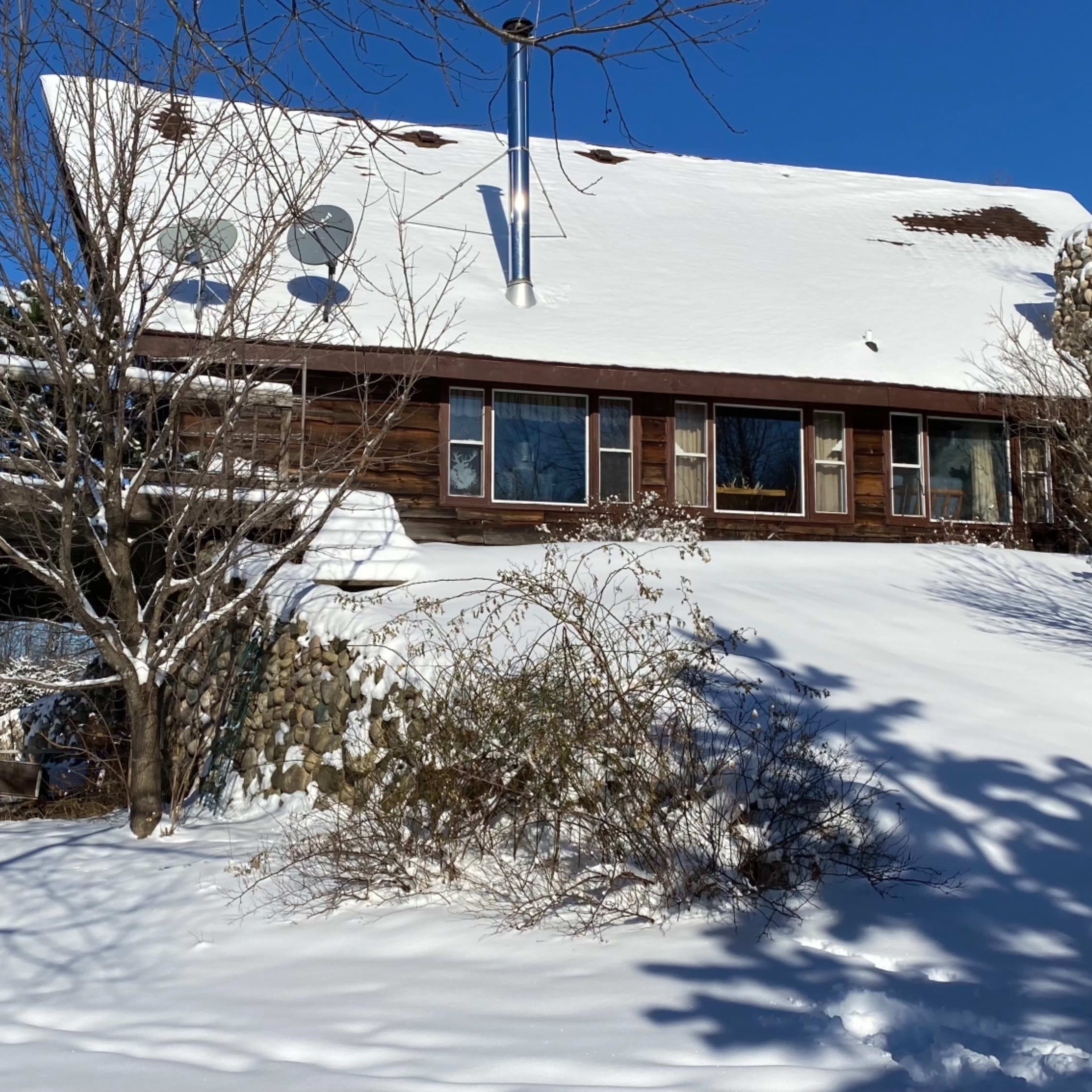 A wooden house with snow-covered roof and surrounding landscape is set on a hillside, partially obscured by trees and bushes, under a clear blue sky.