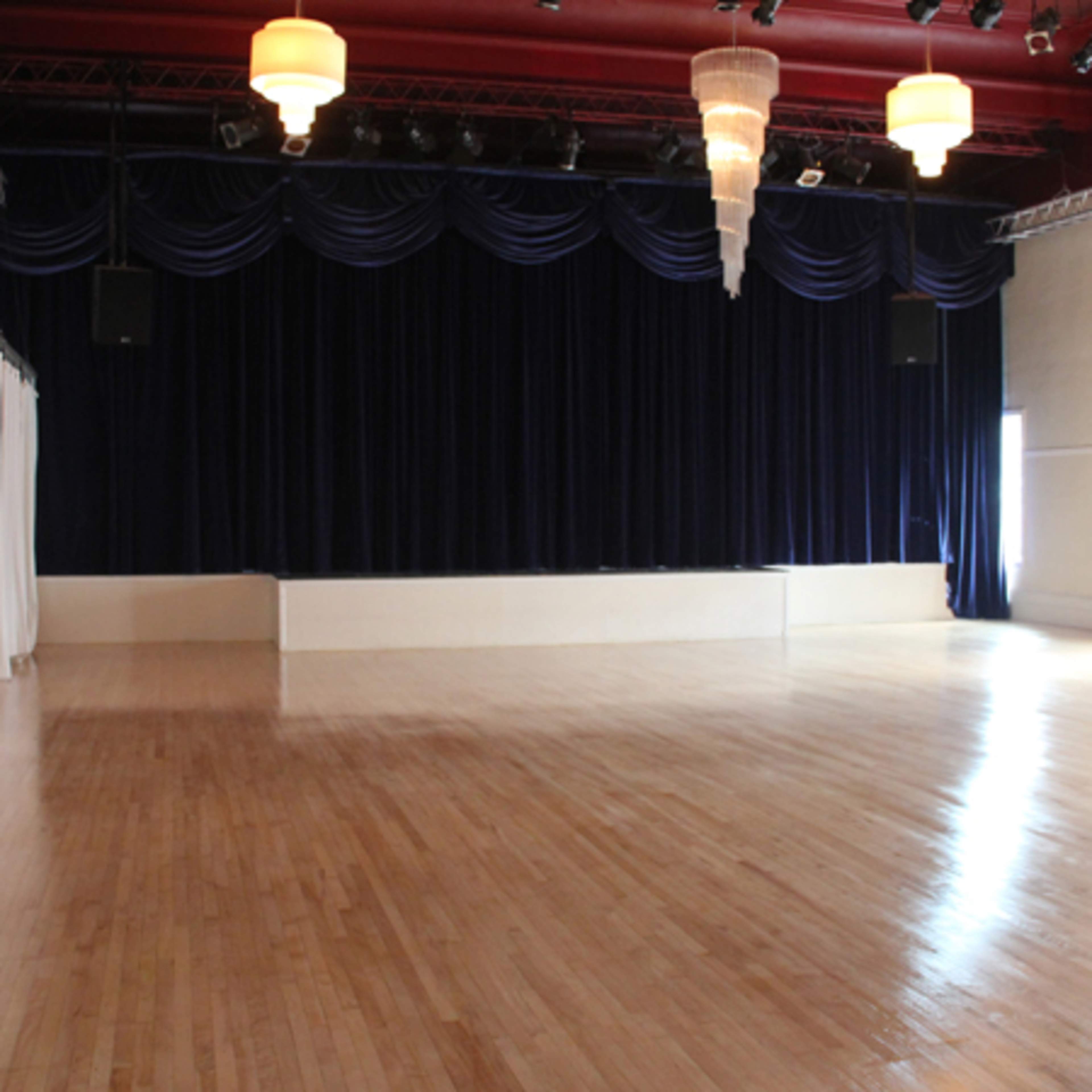 An empty dance hall with polished wooden floors, dark draped curtains, and a hanging chandelier.