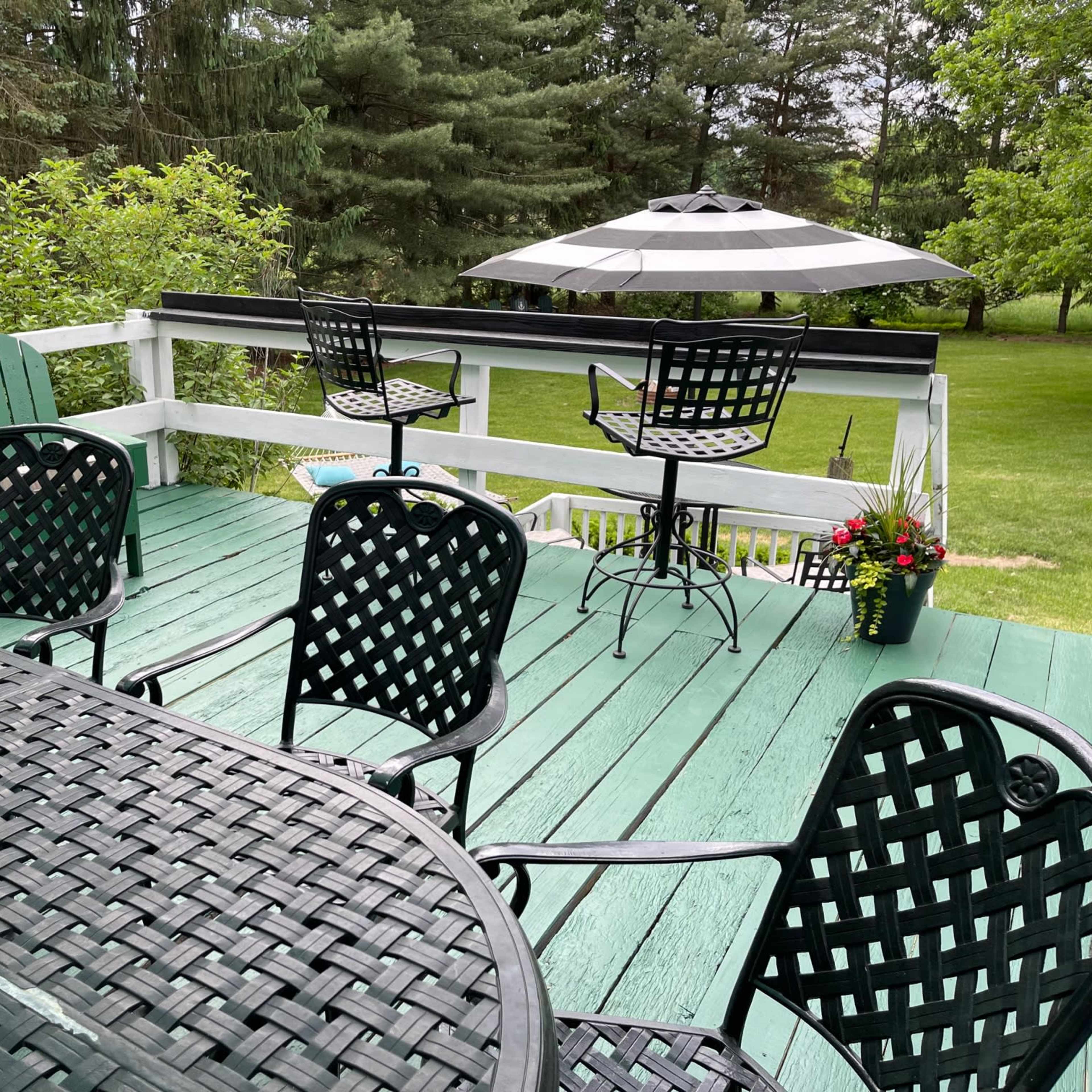 A spacious outdoor deck features black and green chairs, a table with a lantern, and an umbrella over two high-top seats, surrounded by greenery.