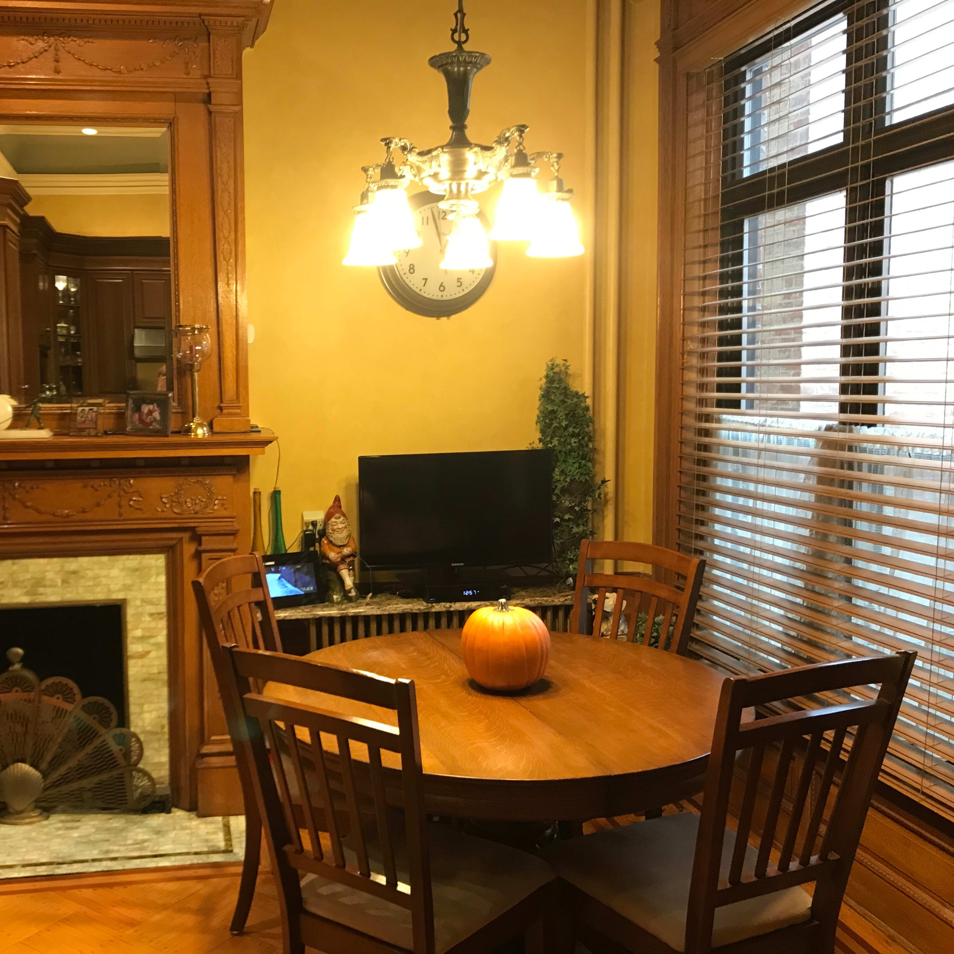 A wooden dining table with four chairs is positioned near a fireplace and a television in a warmly lit room.