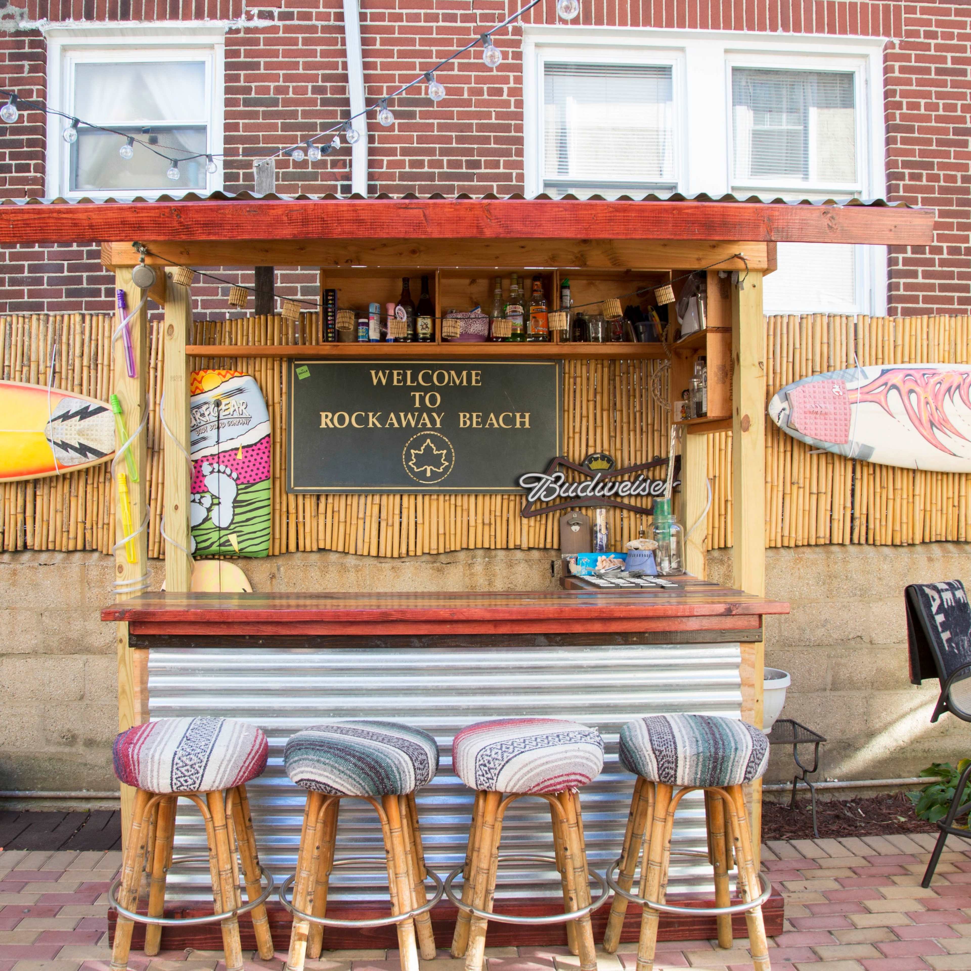 A beach bar with a wooden counter and stools displays surfboards and a sign welcoming visitors to Rockaway Beach.