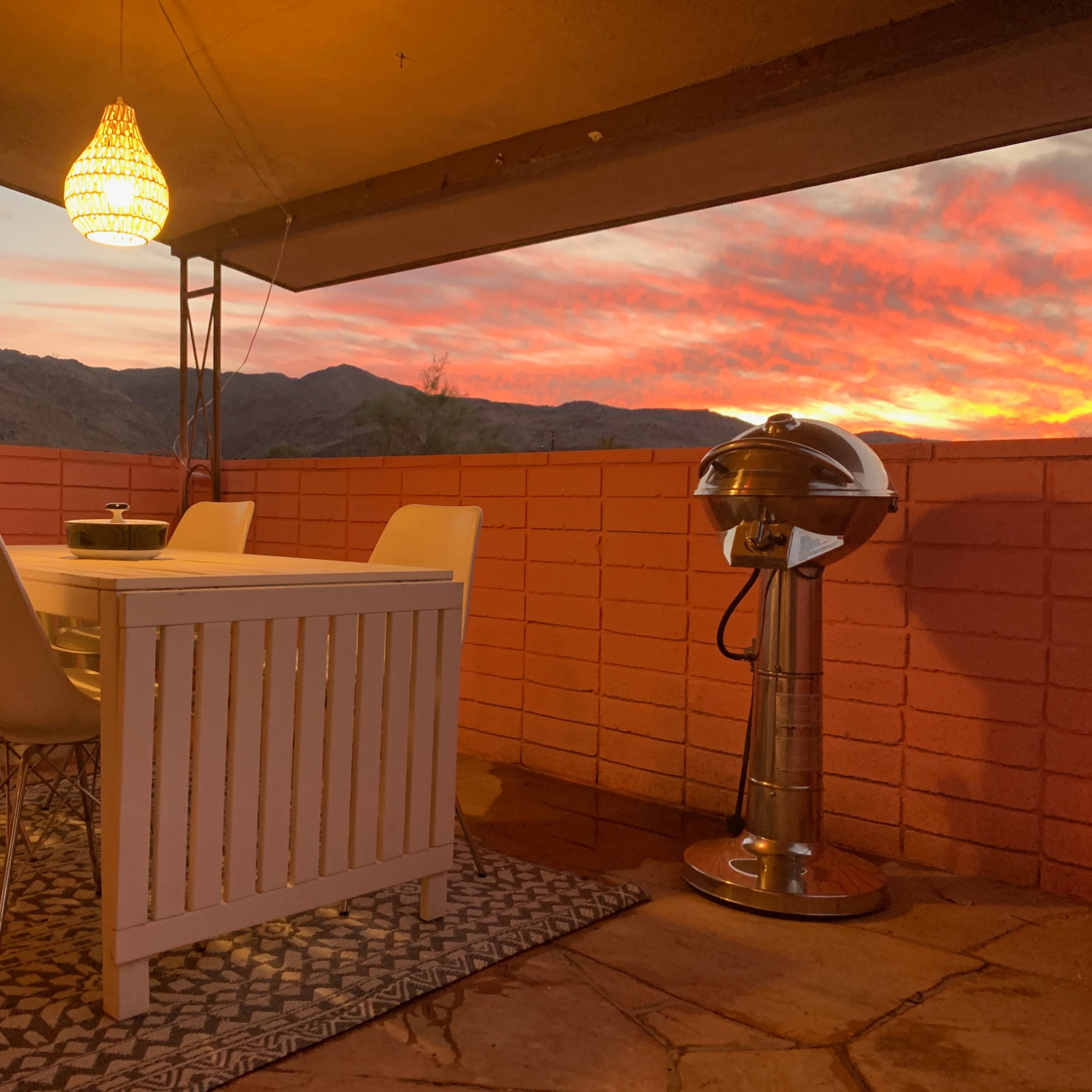 A outdoor dining area features a white table with four chairs and a grill, set against a backdrop of a vibrant sunset over the mountains.
