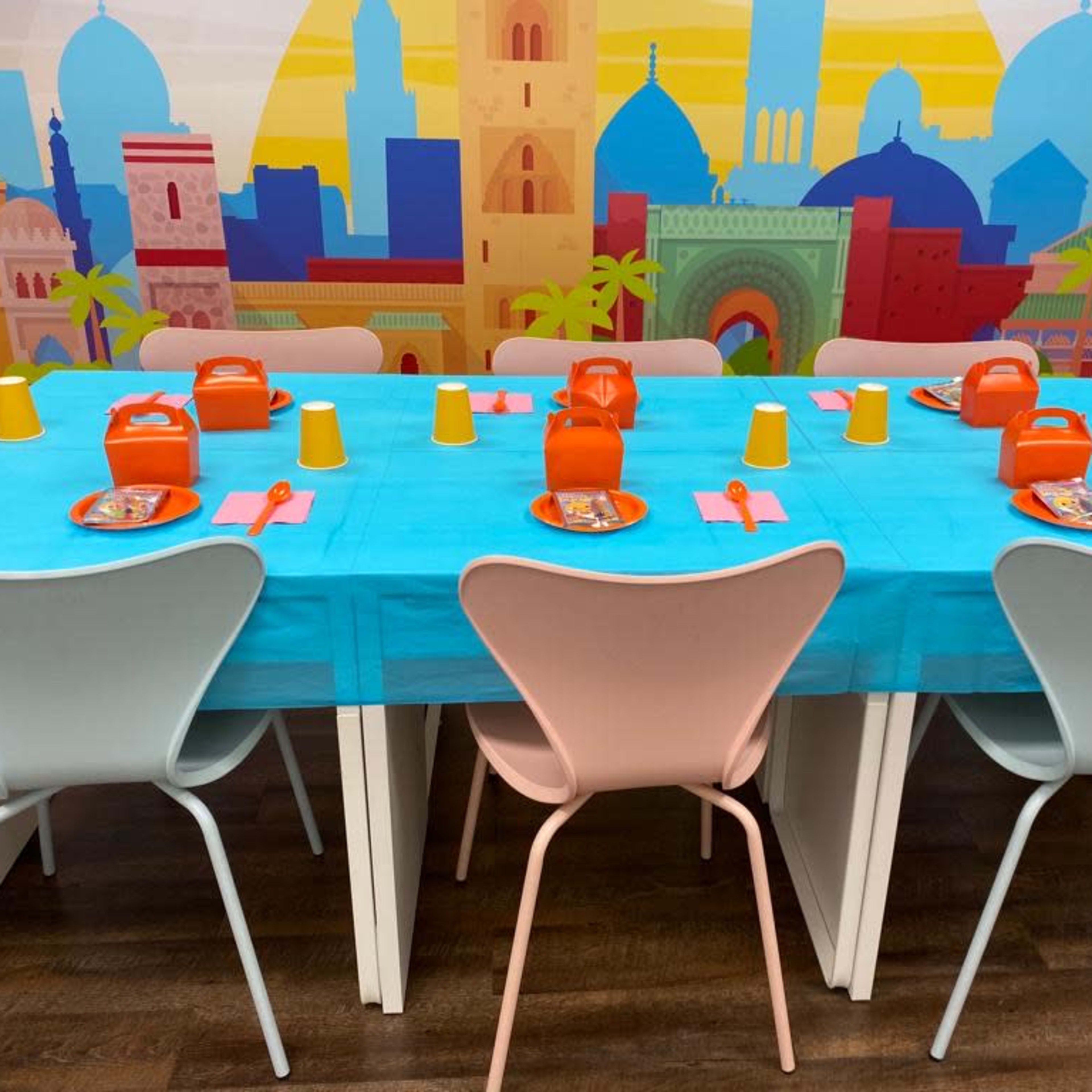 A dining table is set with blue tablecloths, orange plates, yellow cups, and pink utensils, against a colorful background featuring cityscapes and palm trees.