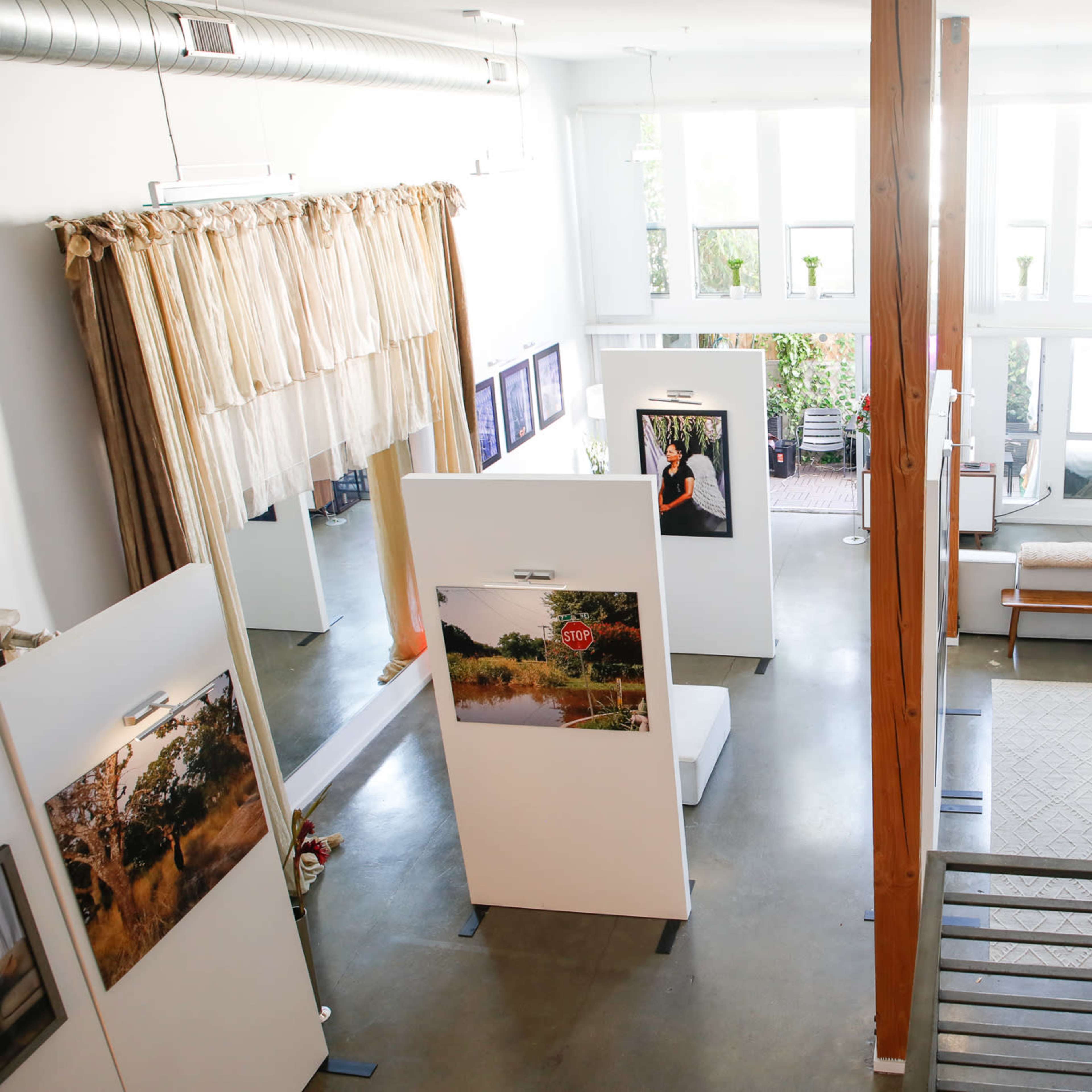 A spacious art gallery with white walls, tall windows, and exhibition panels displaying photographs surrounded by a modern seating area.