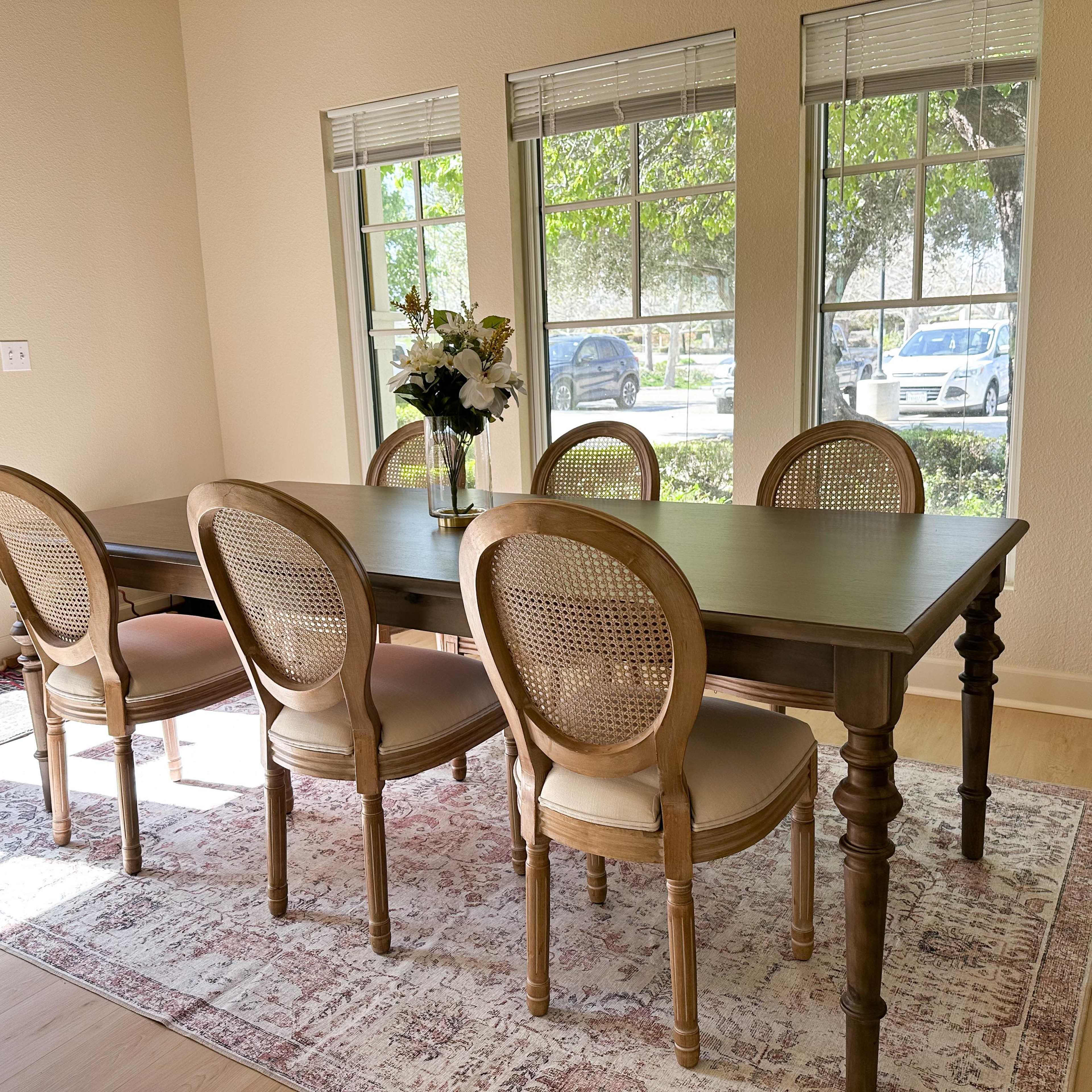 A rectangular dining table with six cane-backed chairs is positioned under large windows in a brightly lit room.