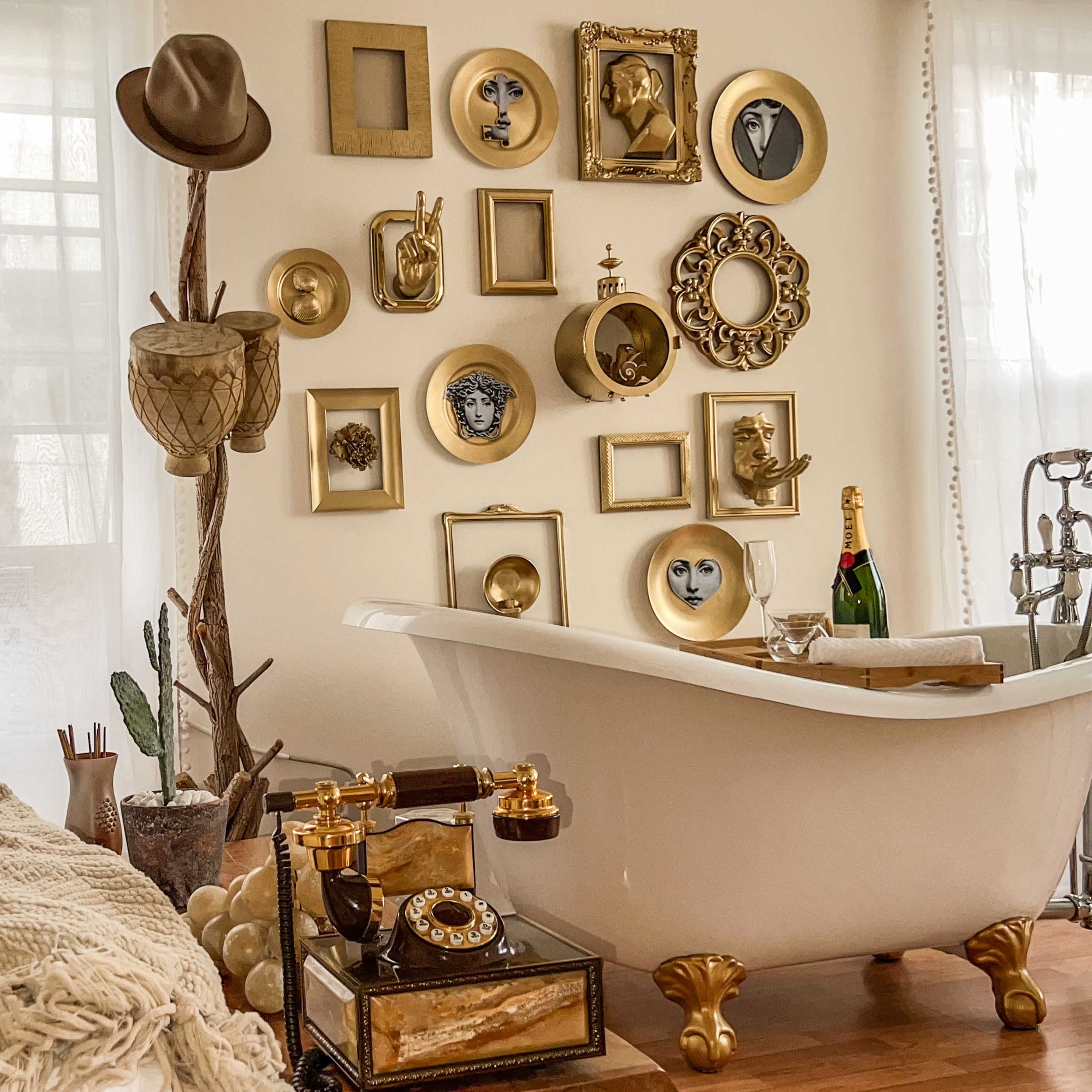 A vintage-style bathroom featuring a freestanding bathtub, decorative wall art in gold frames, and a small side table with a vintage telephone and champagne.