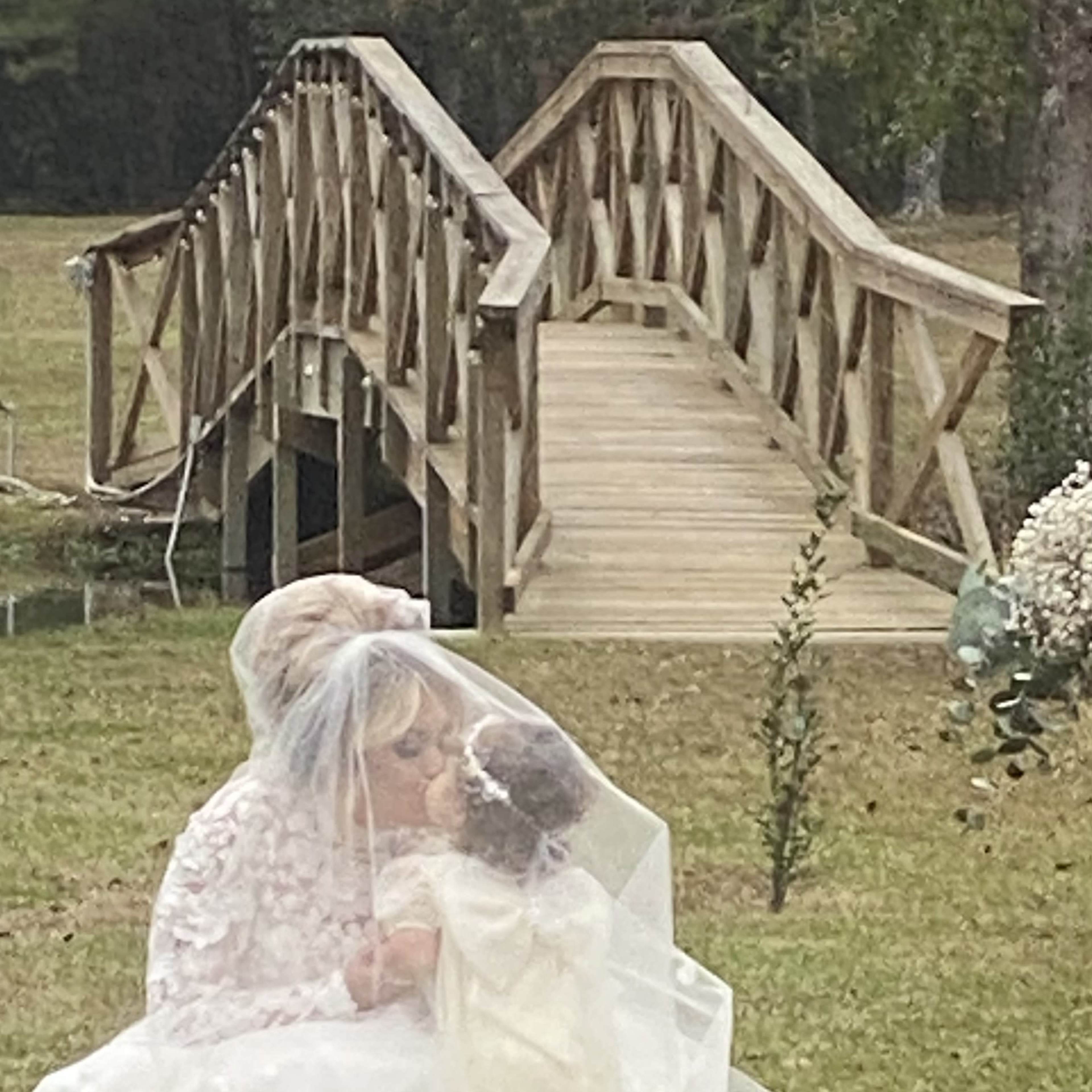 A woman in a wedding dress kneels to embrace a young girl in a white outfit near a wooden bridge in a grassy area.