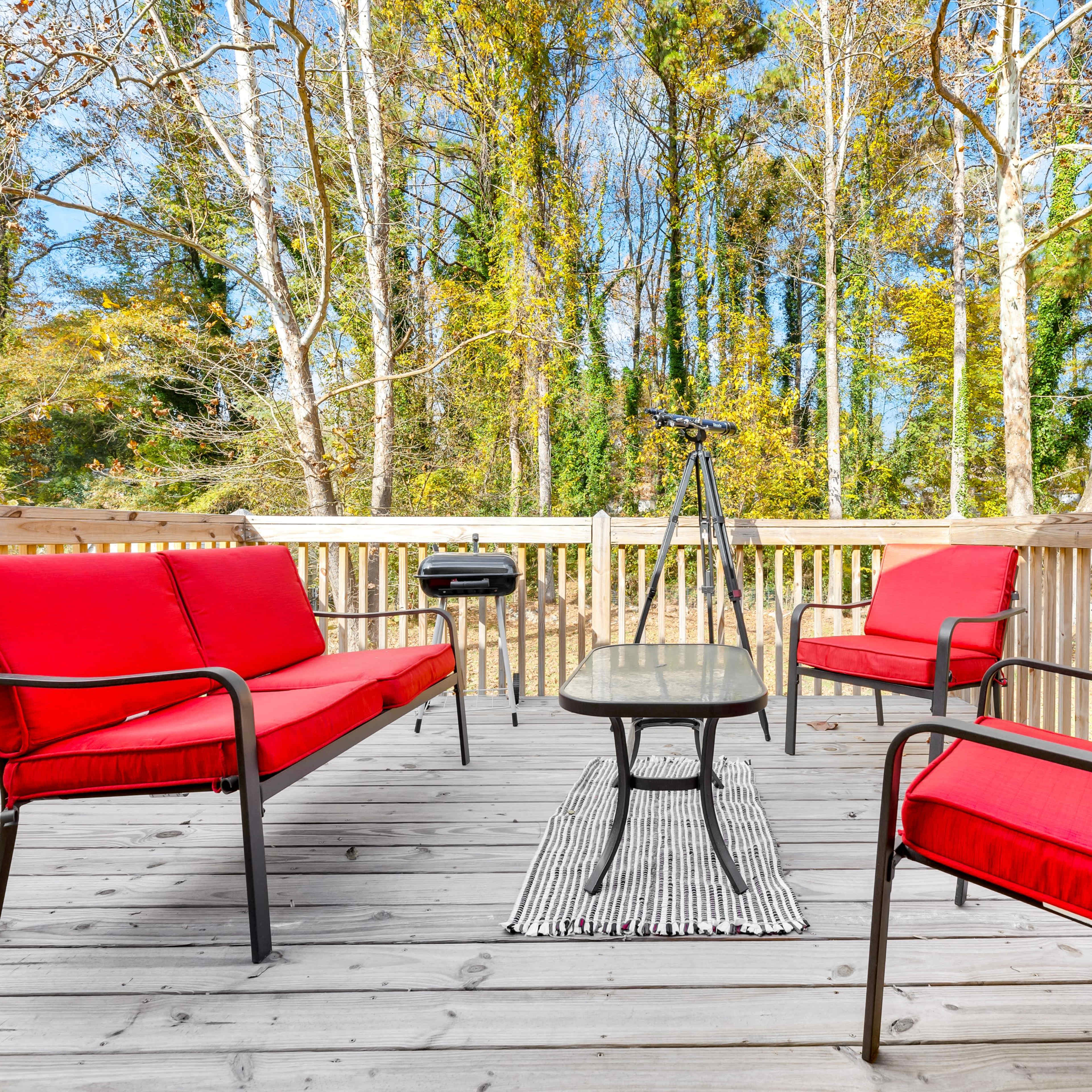 A wooden deck features red chairs and a table, surrounded by trees and a grill in the background.