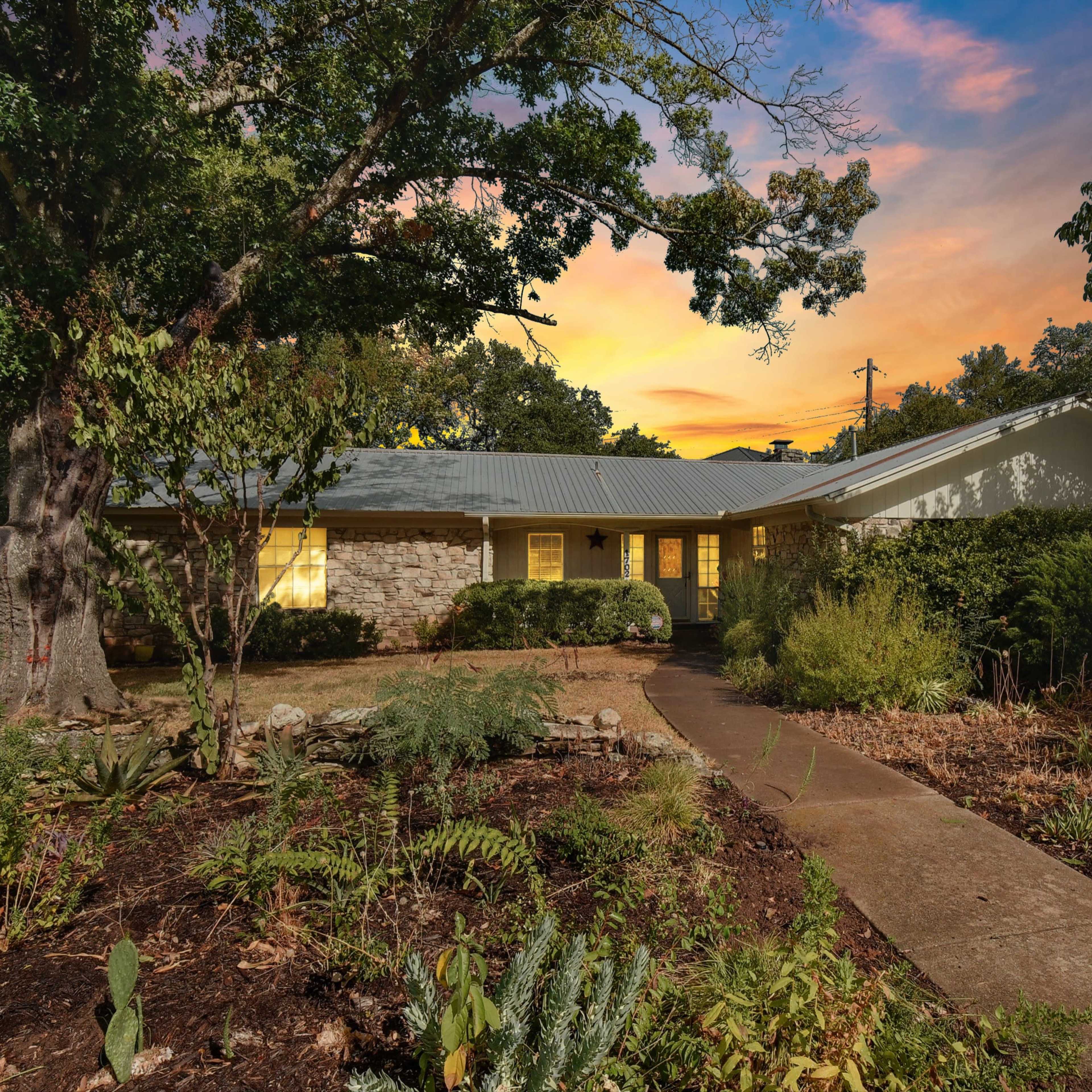 A single-story house with a stone facade, surrounded by a landscaped yard featuring various plants and trees under a sunset sky.