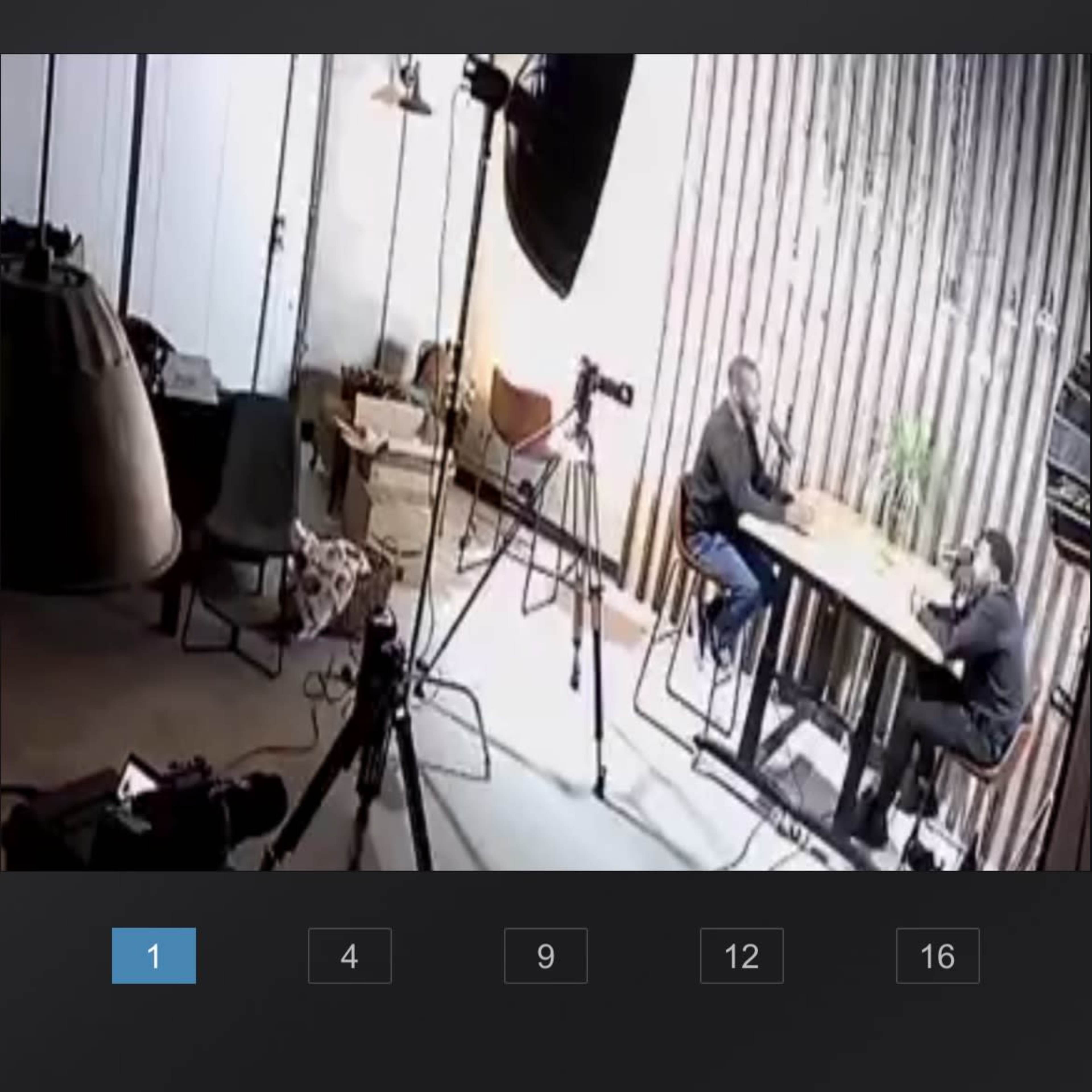 A camera captures two people sitting at a table in an office setting, surrounded by equipment and furniture.