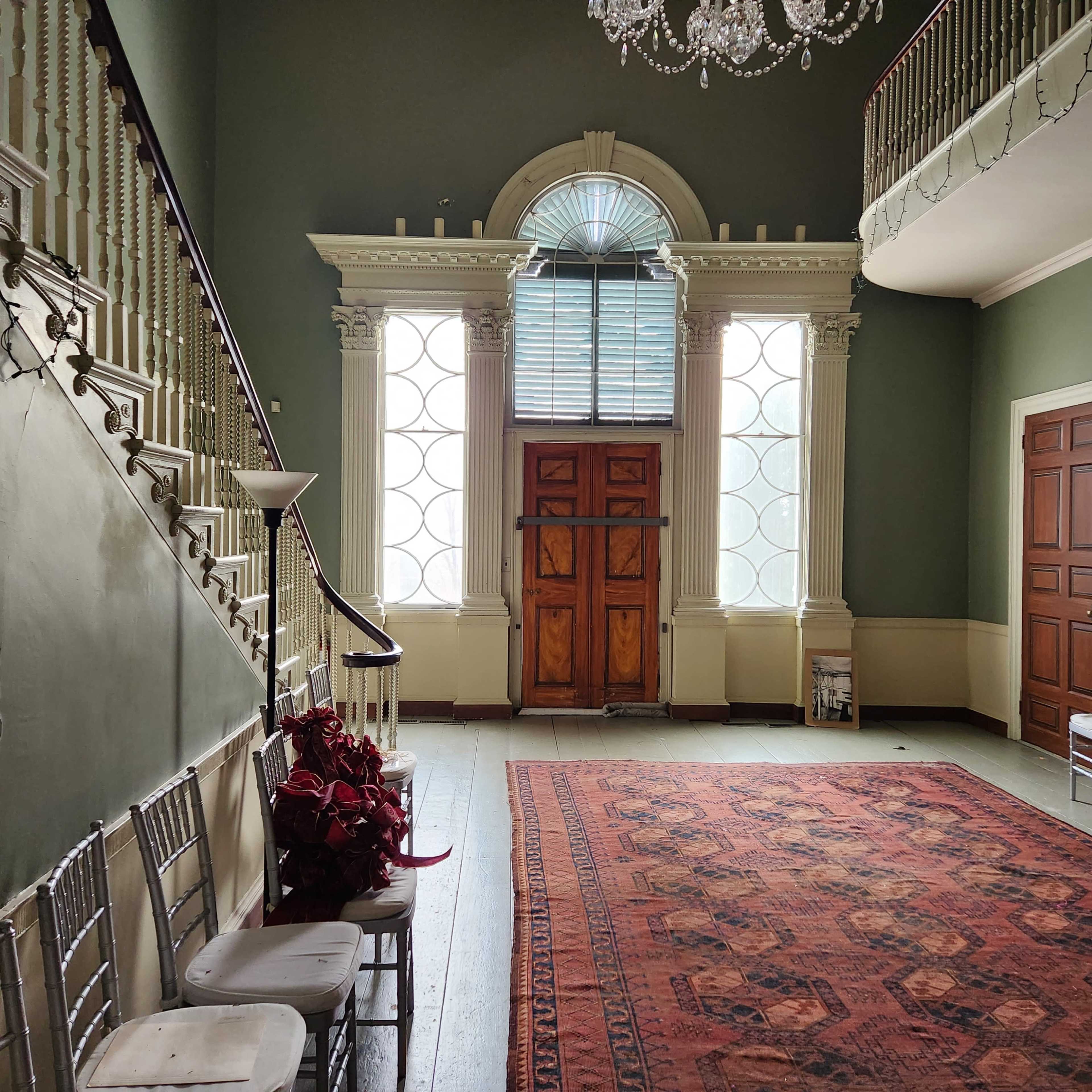 A spacious foyer with a large rug, a staircase on the left, elegant chairs, and a prominent chandelier above, flanked by tall windows and a wooden door.