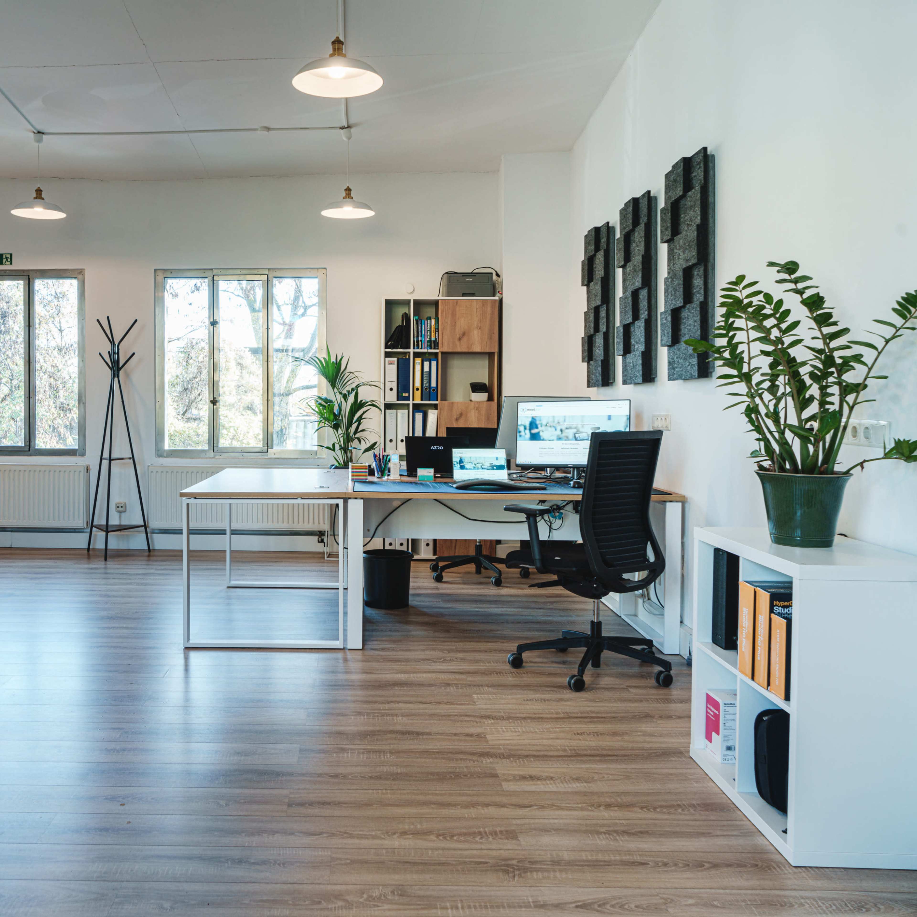 A modern office space with a desk, computer, and plants, featuring large windows and wooden flooring.