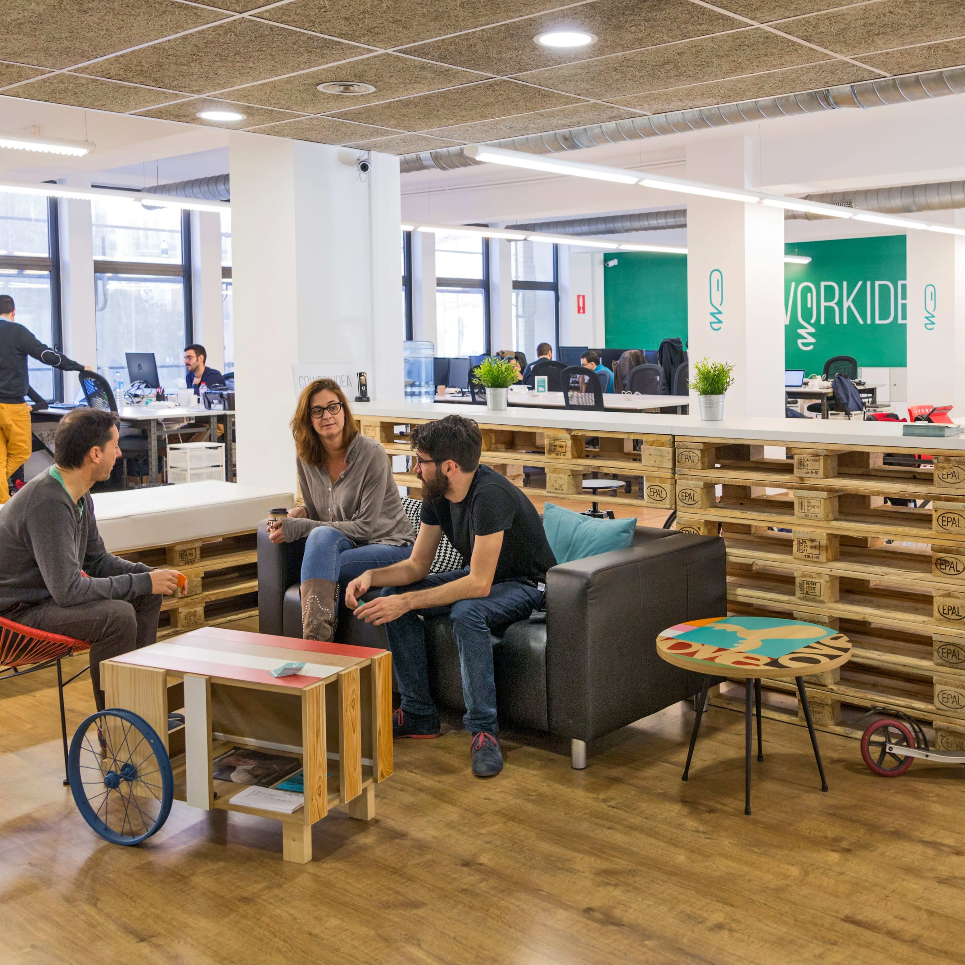 A modern office space features seating areas with pallet furniture, a scooter, and several workers engaged at desks in the background.