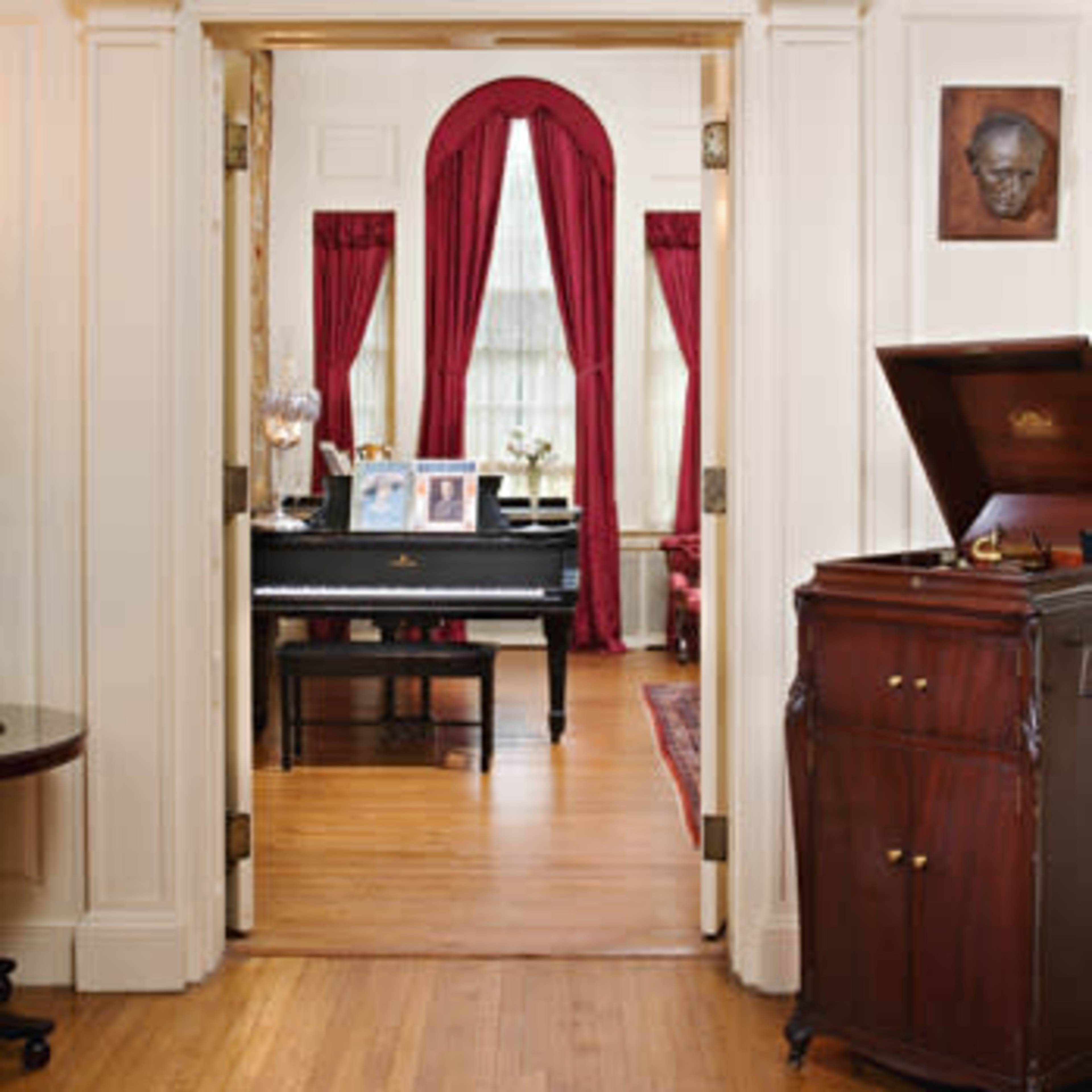 A doorway framed by decorative molding leads into a room with a piano, photographs on display, and rich red curtains.