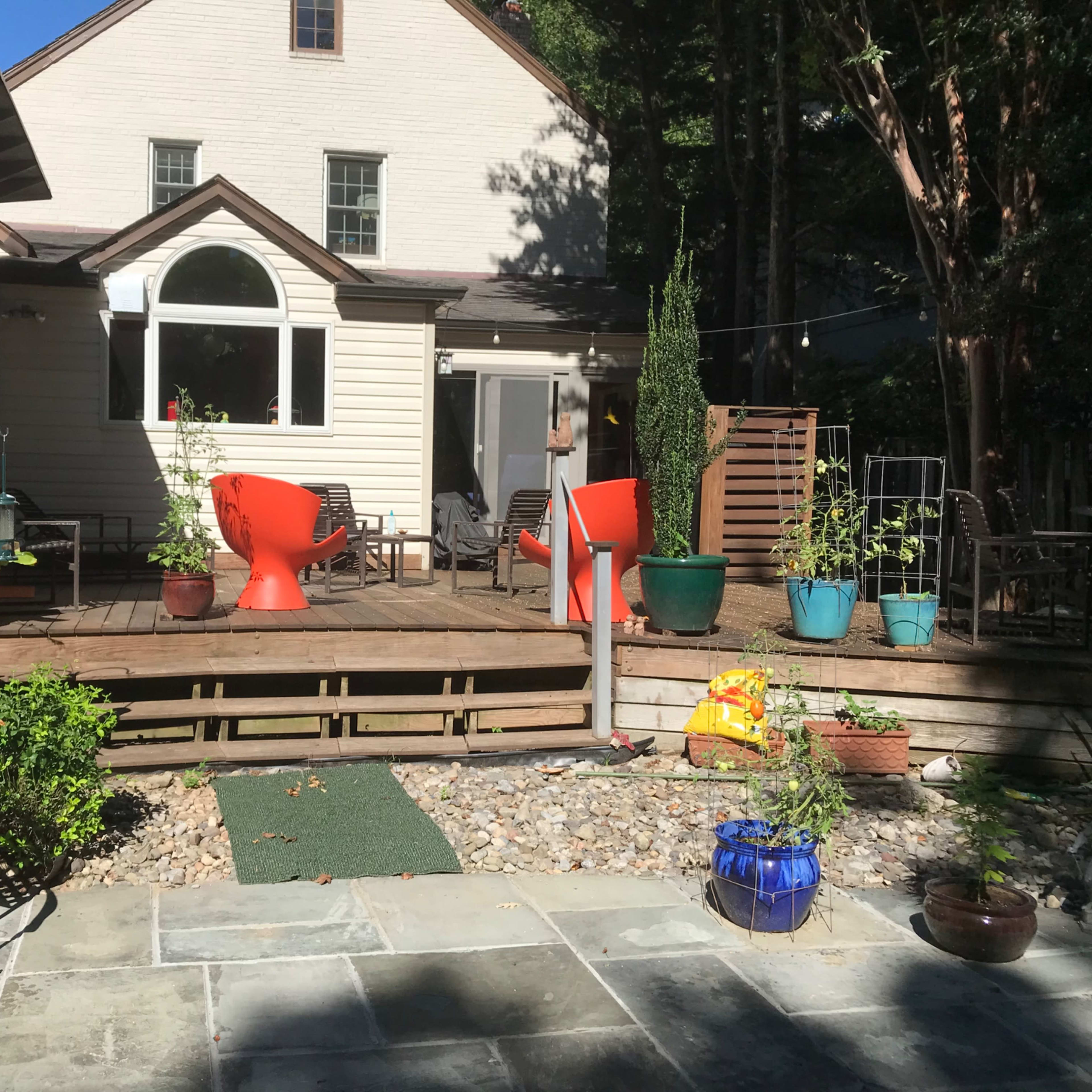 A backyard patio area with a wooden deck, two large red chairs, potted plants, and a stone pathway.