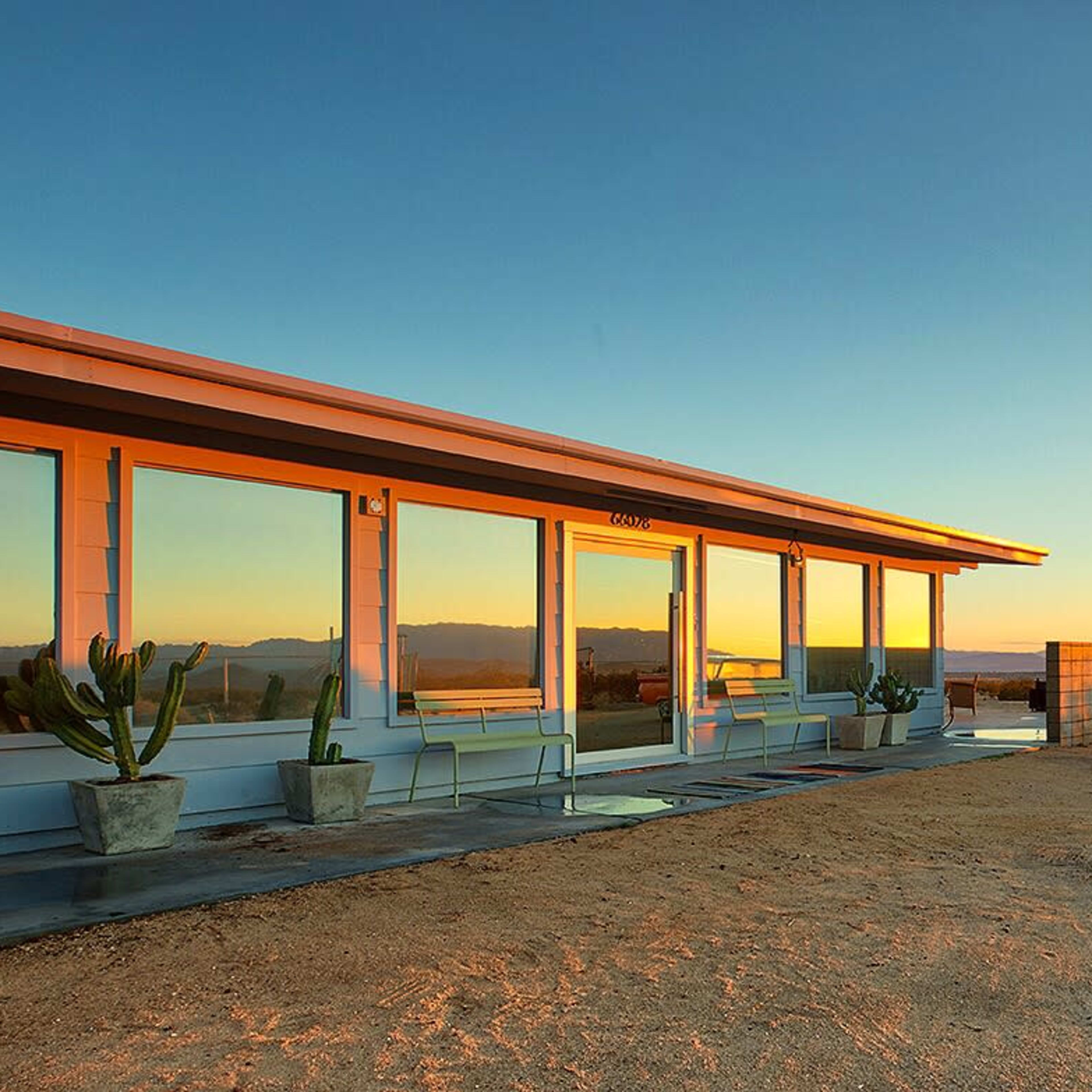 A modern building with large windows sits on a desert landscape, accompanied by a vintage car parked nearby at sunset.