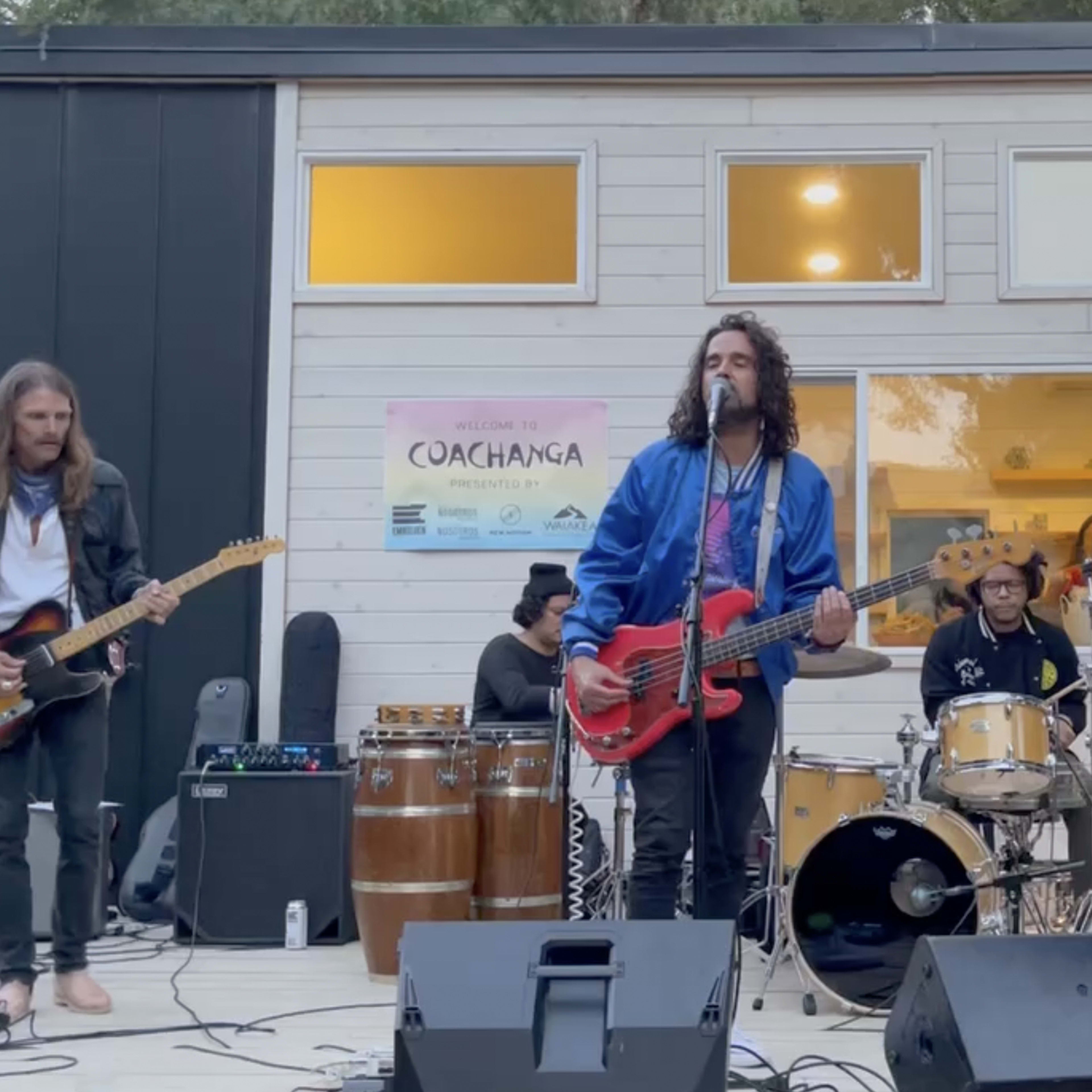 A band performs on an outdoor stage with various instruments, including electric guitars, drums, and a keyboard.