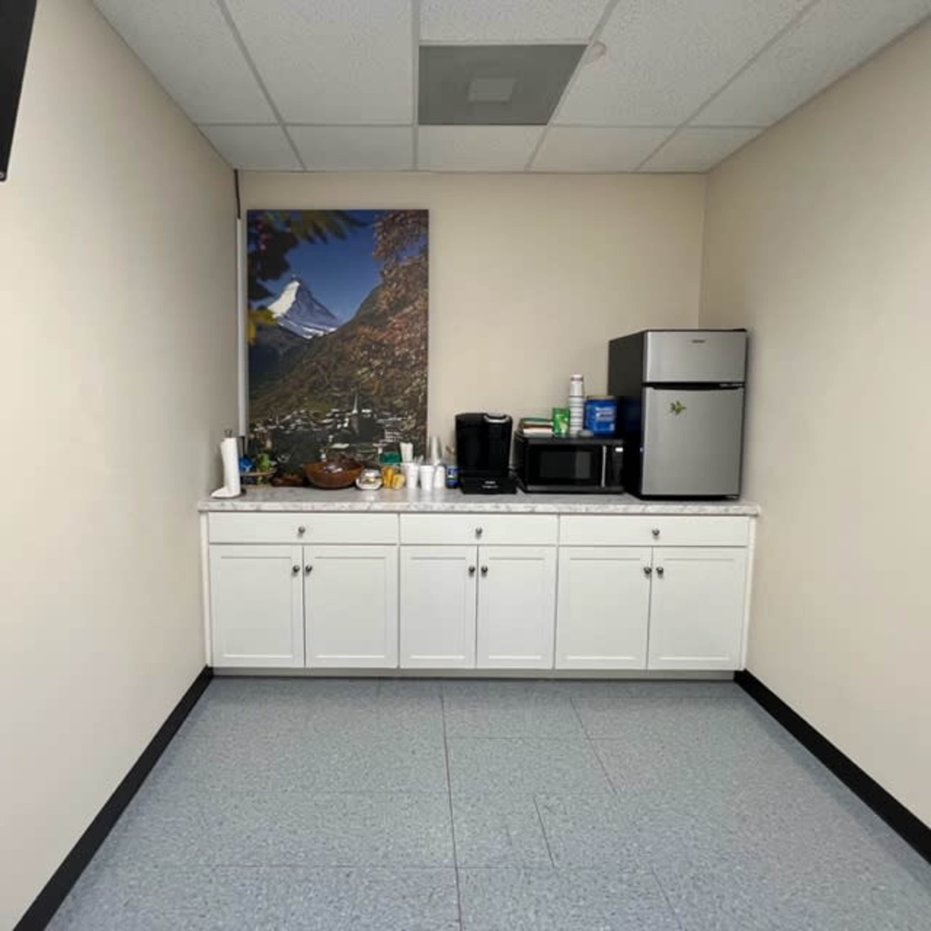 A break room with a white cabinet, a small refrigerator, a coffee maker, a microwave, and a scenic mountain painting on the wall.