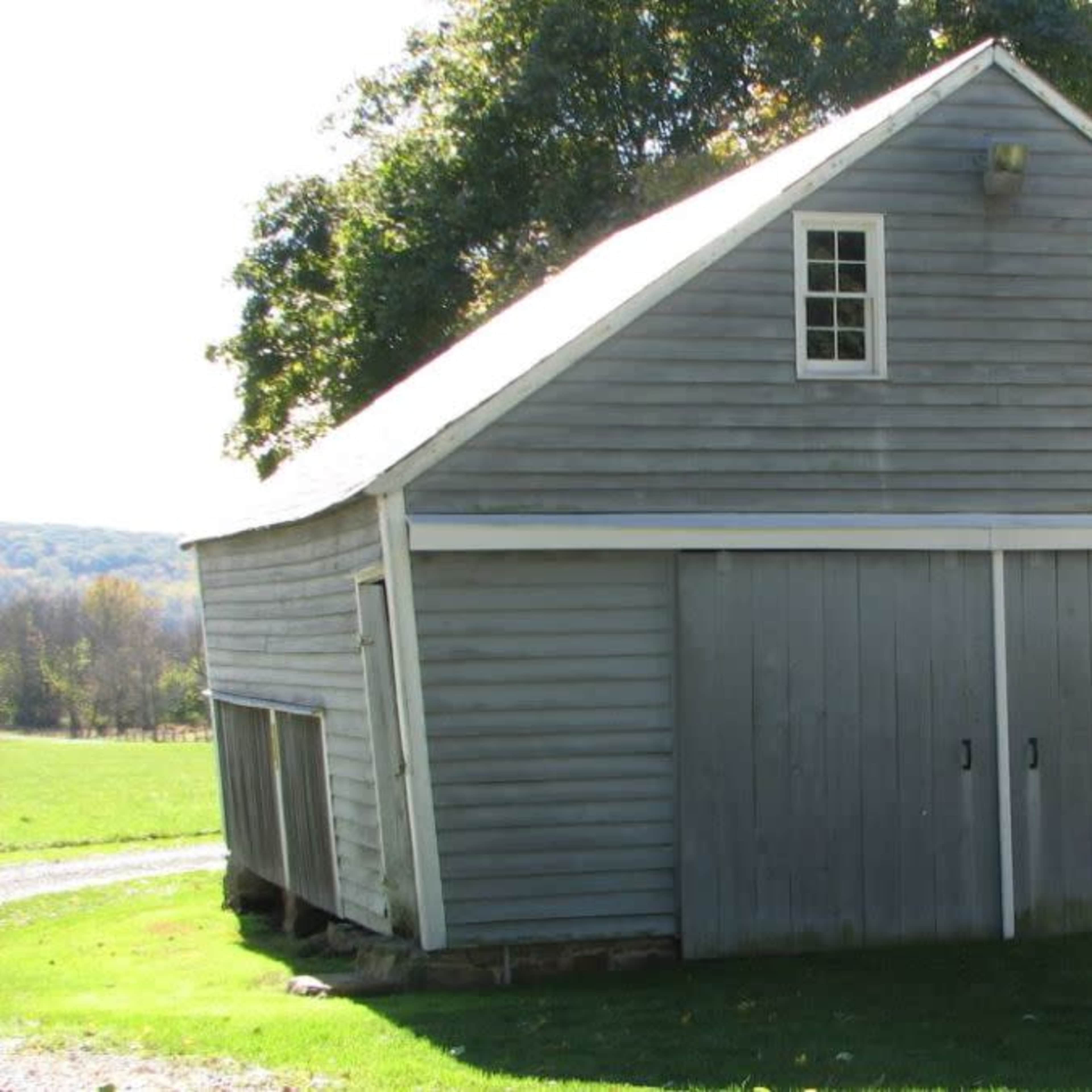 A gray wooden barn with two doors, set on a grassy area beside a gravel path.