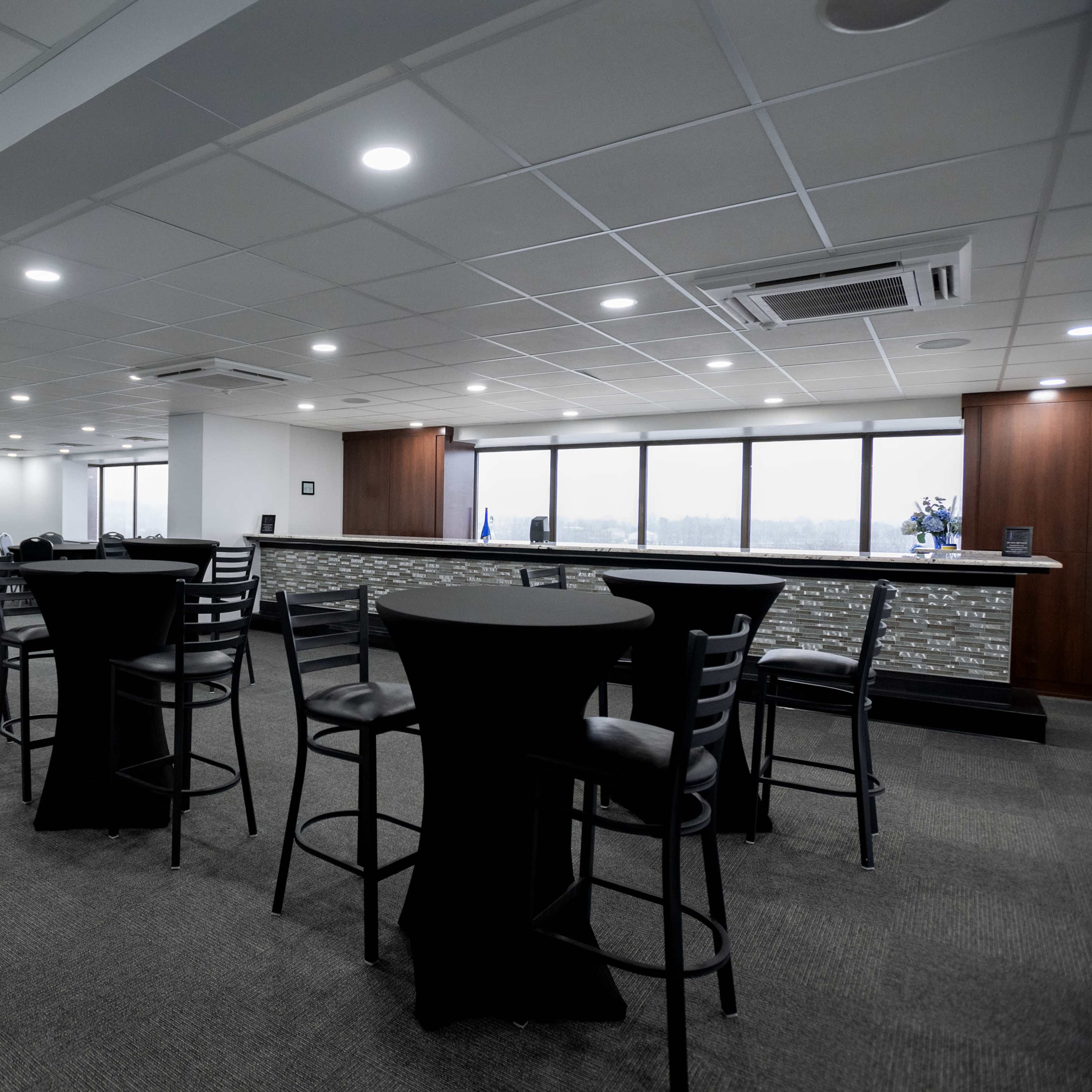 A modern office lounge area with black high-top tables and chairs, well-lit by ceiling lights and natural light from large windows.