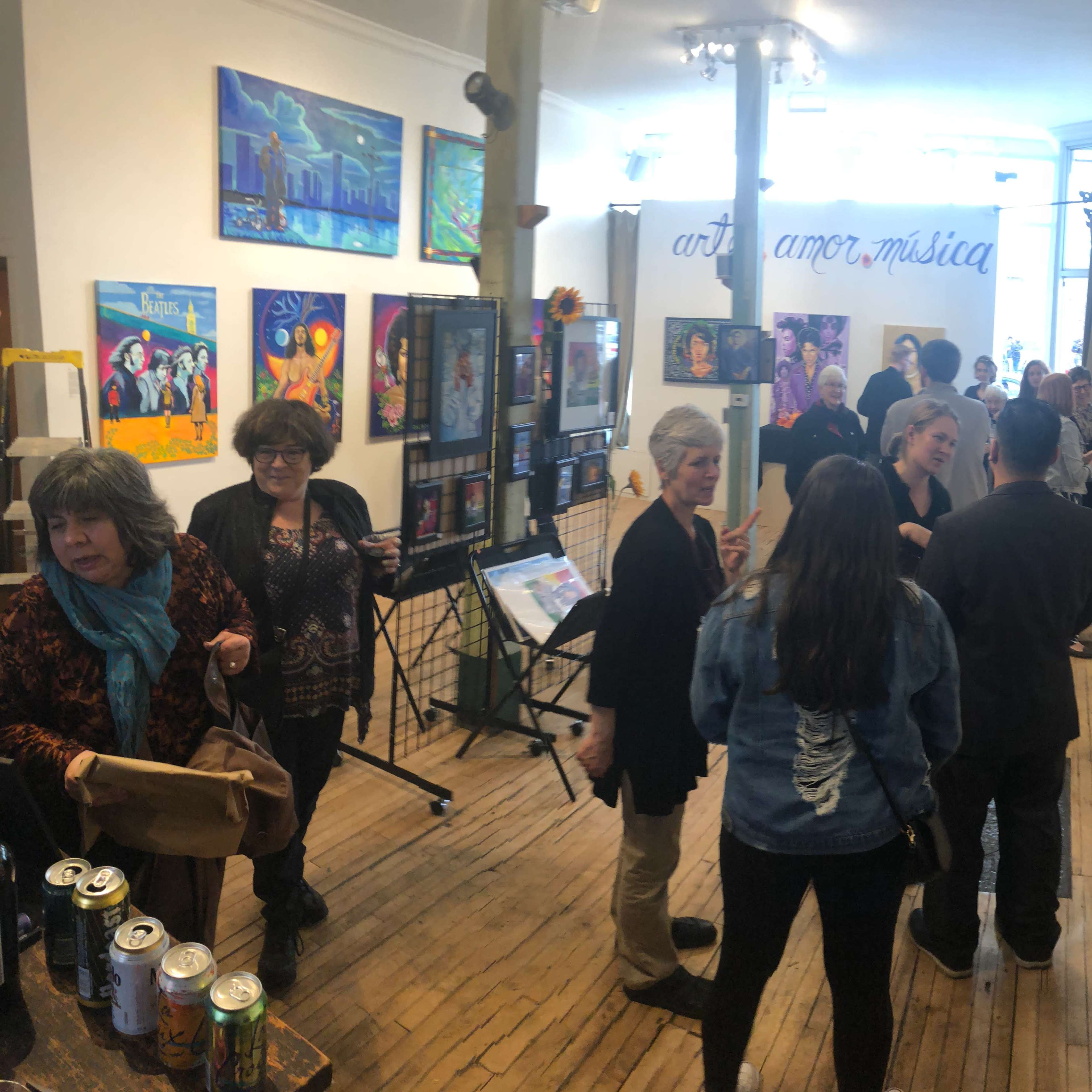 A group of people gathers in an art gallery featuring colorful paintings and a display of various artworks along the walls.
