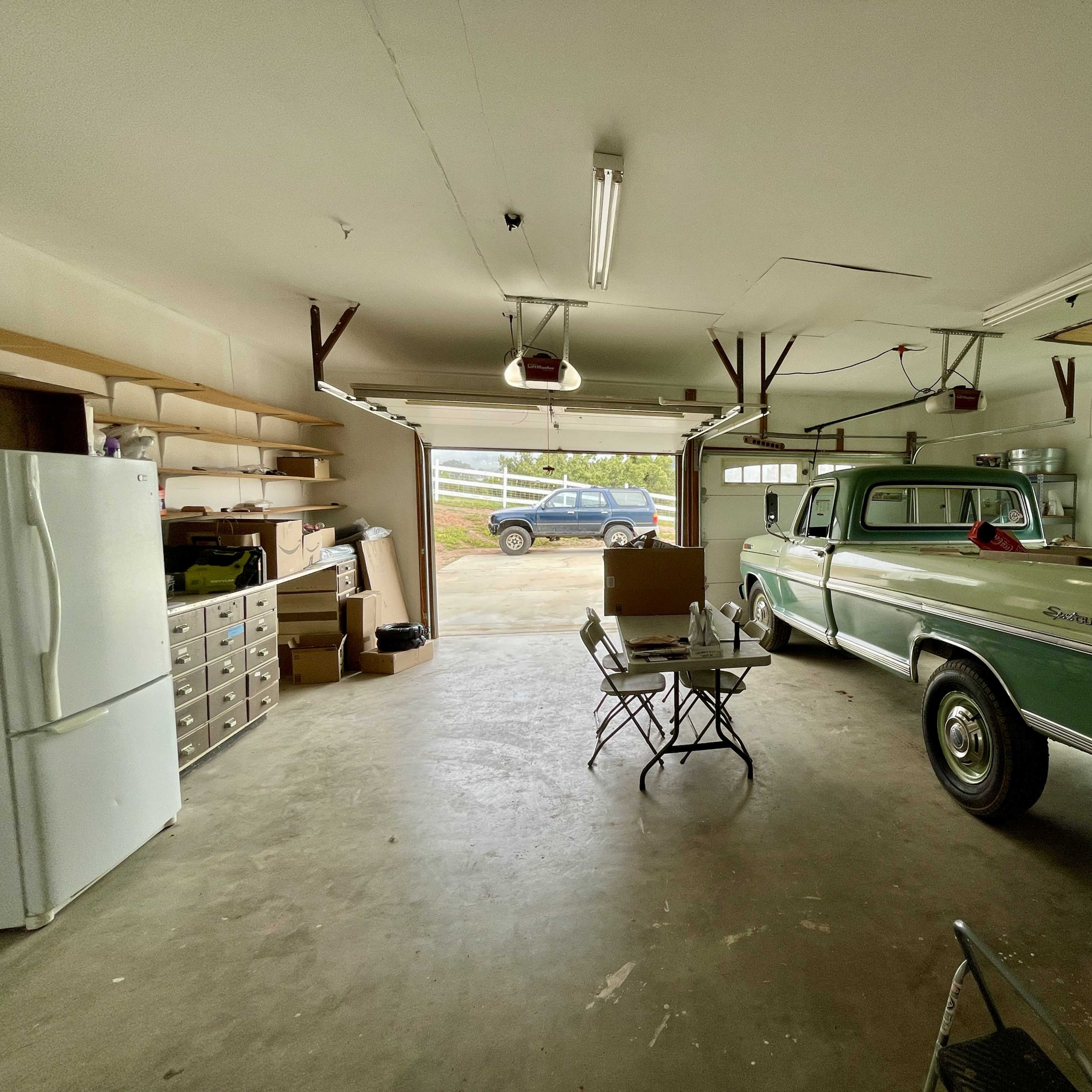 A green Ford pickup truck is parked inside a spacious garage with storage shelves and a refrigerator visible along the walls.