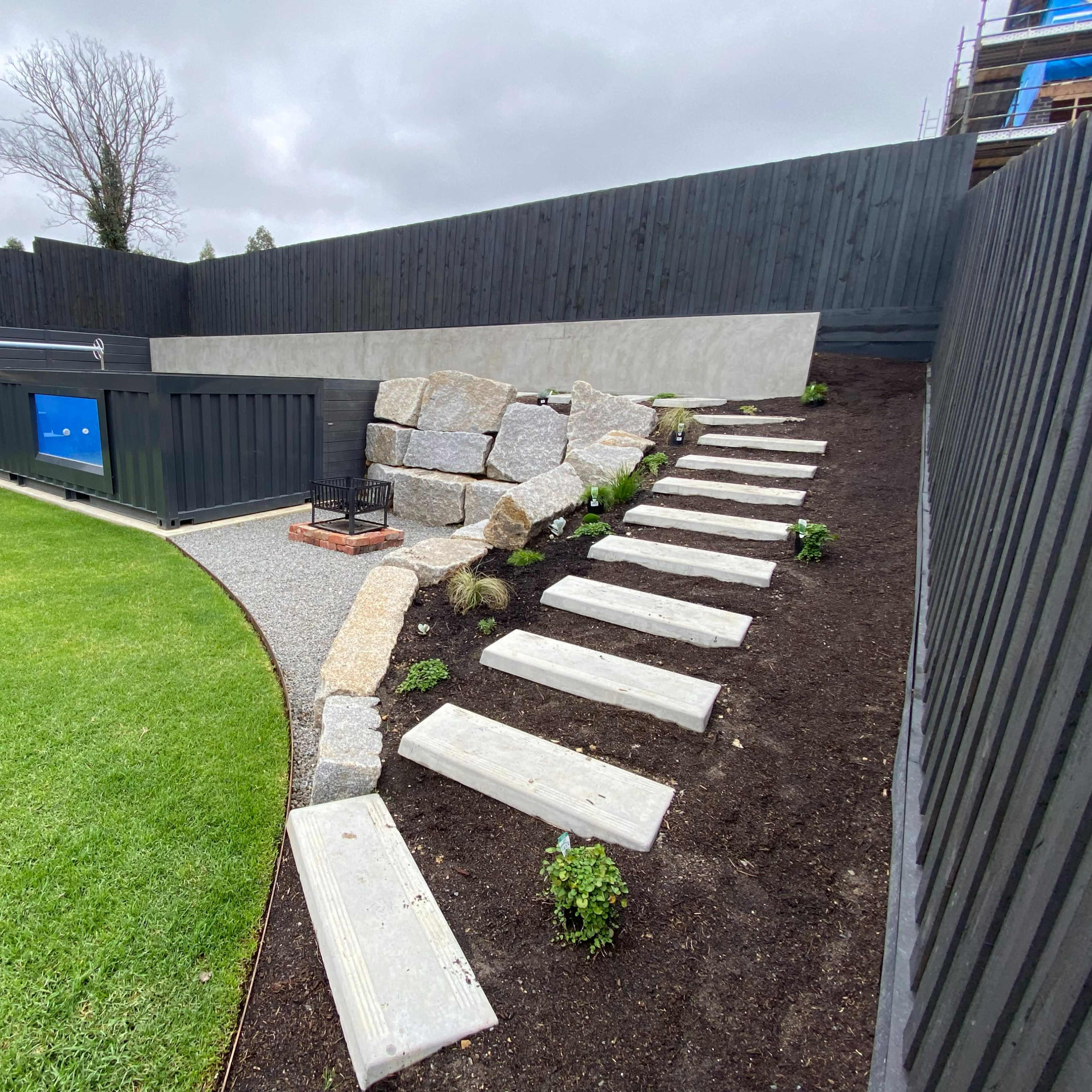 A landscaped yard featuring a grassy area, a gravel path with stone steps, and a retaining wall with rocks and a container structure.