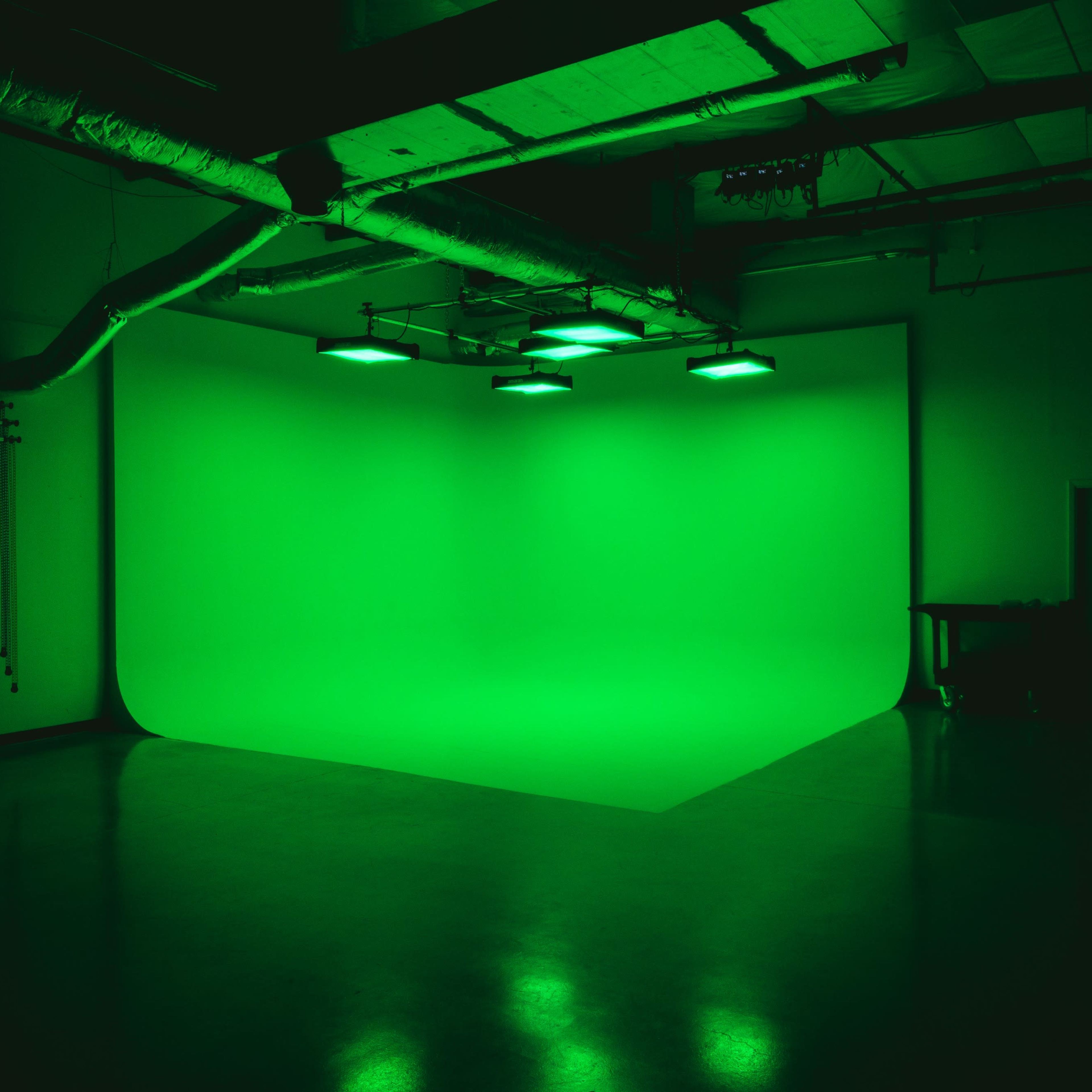 A dimly lit studio features a large green backdrop illuminated by overhead lights.