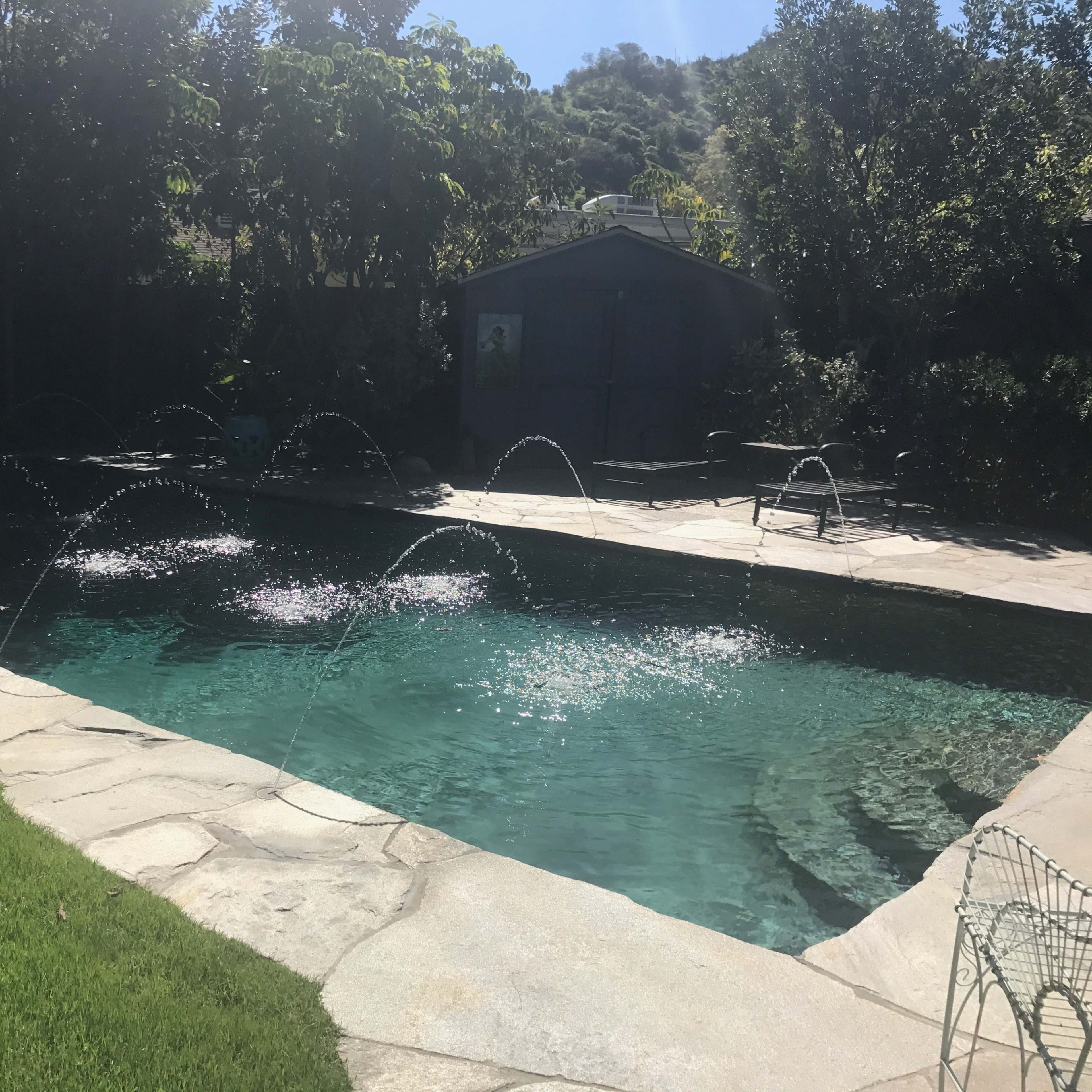 A swimming pool with water fountains sits beside a stone patio and lush greenery.