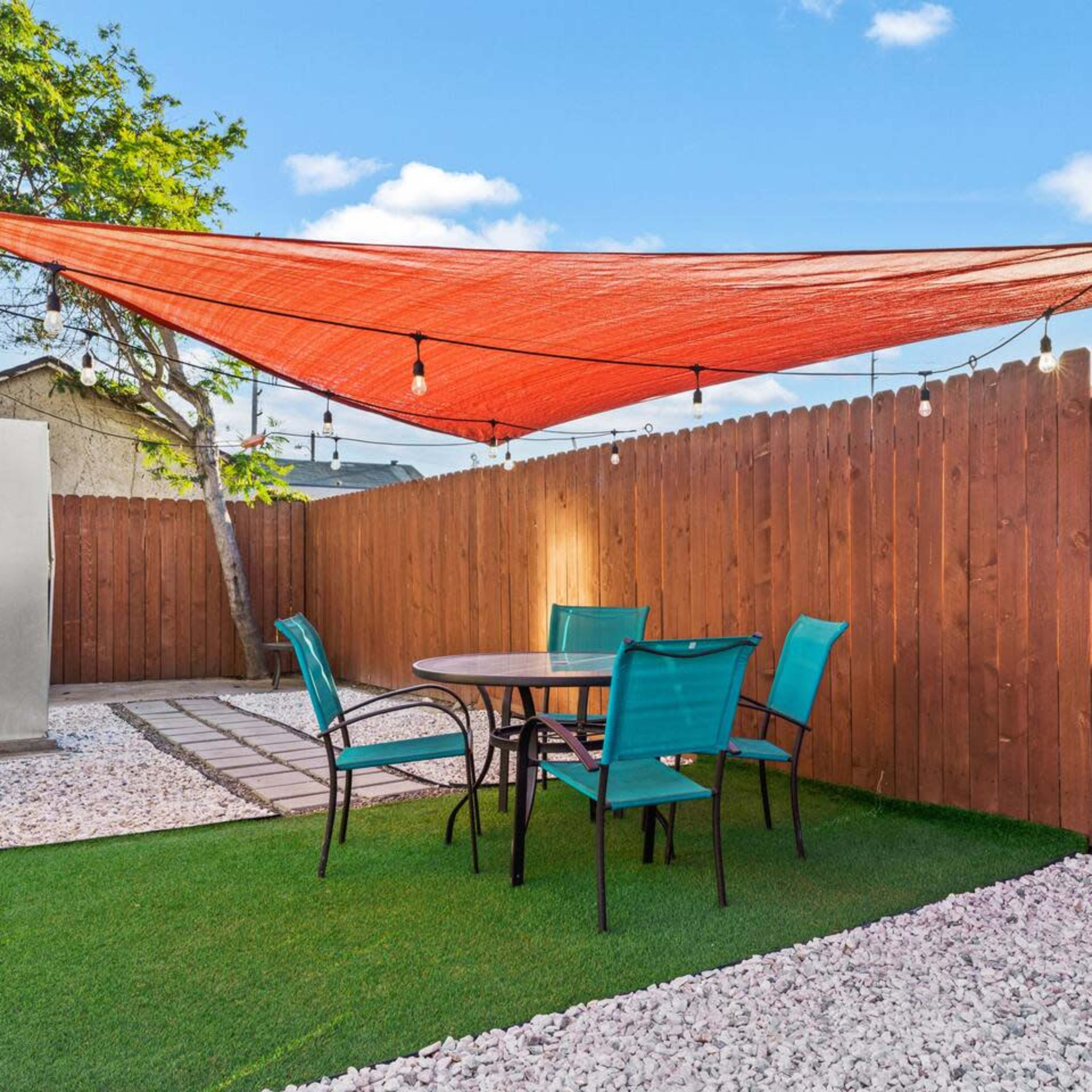 A patio area features a circular table with four turquoise chairs under a red shade sail, surrounded by a gravel and grass landscape.