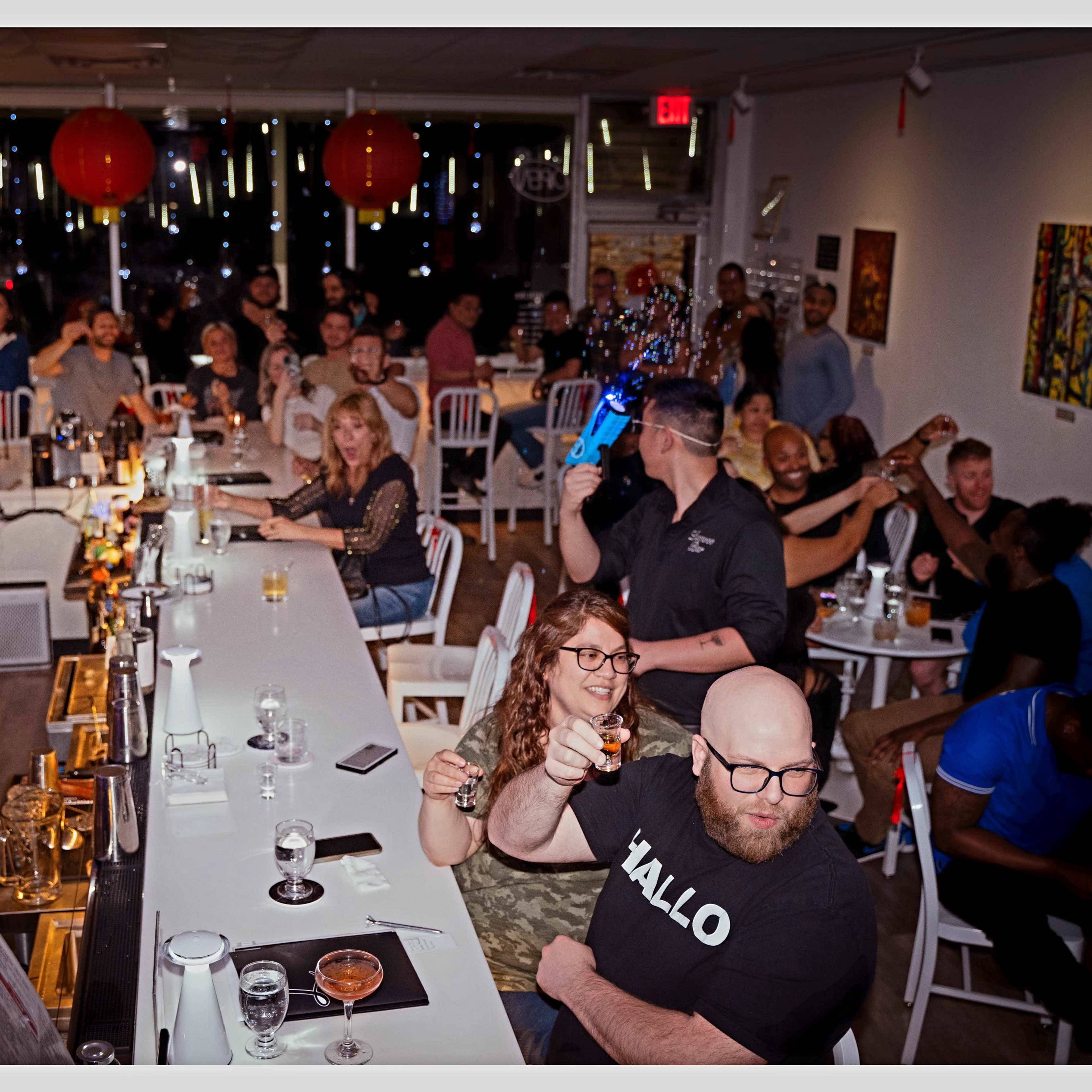 A crowded bar scene shows a mix of patrons enjoying drinks while a bartender serves at a long countertop under dim lighting and decorative elements.