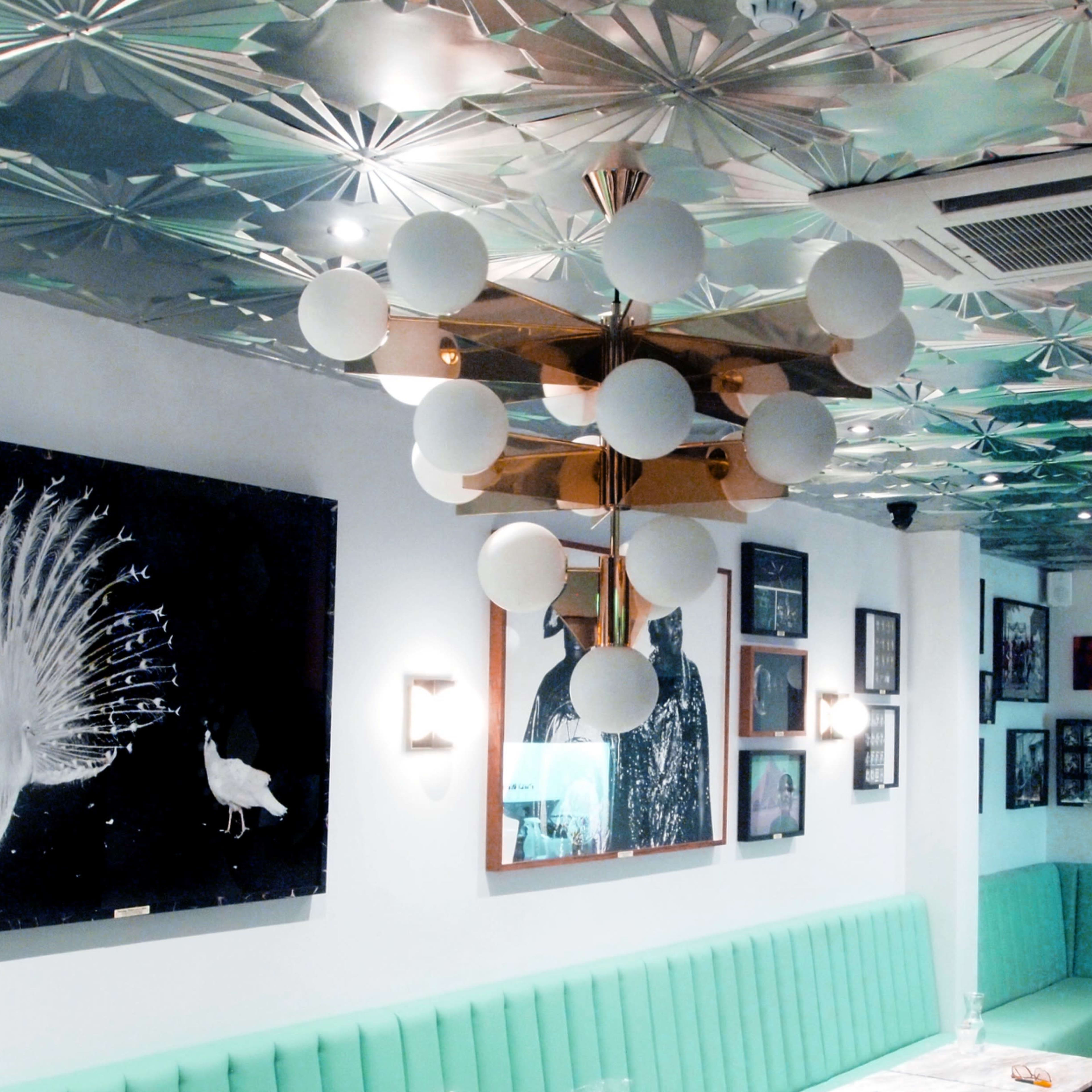 A modern restaurant interior featuring a green banquette, decorative wall art, and a distinctive ceiling with geometric patterns and pendant lighting.