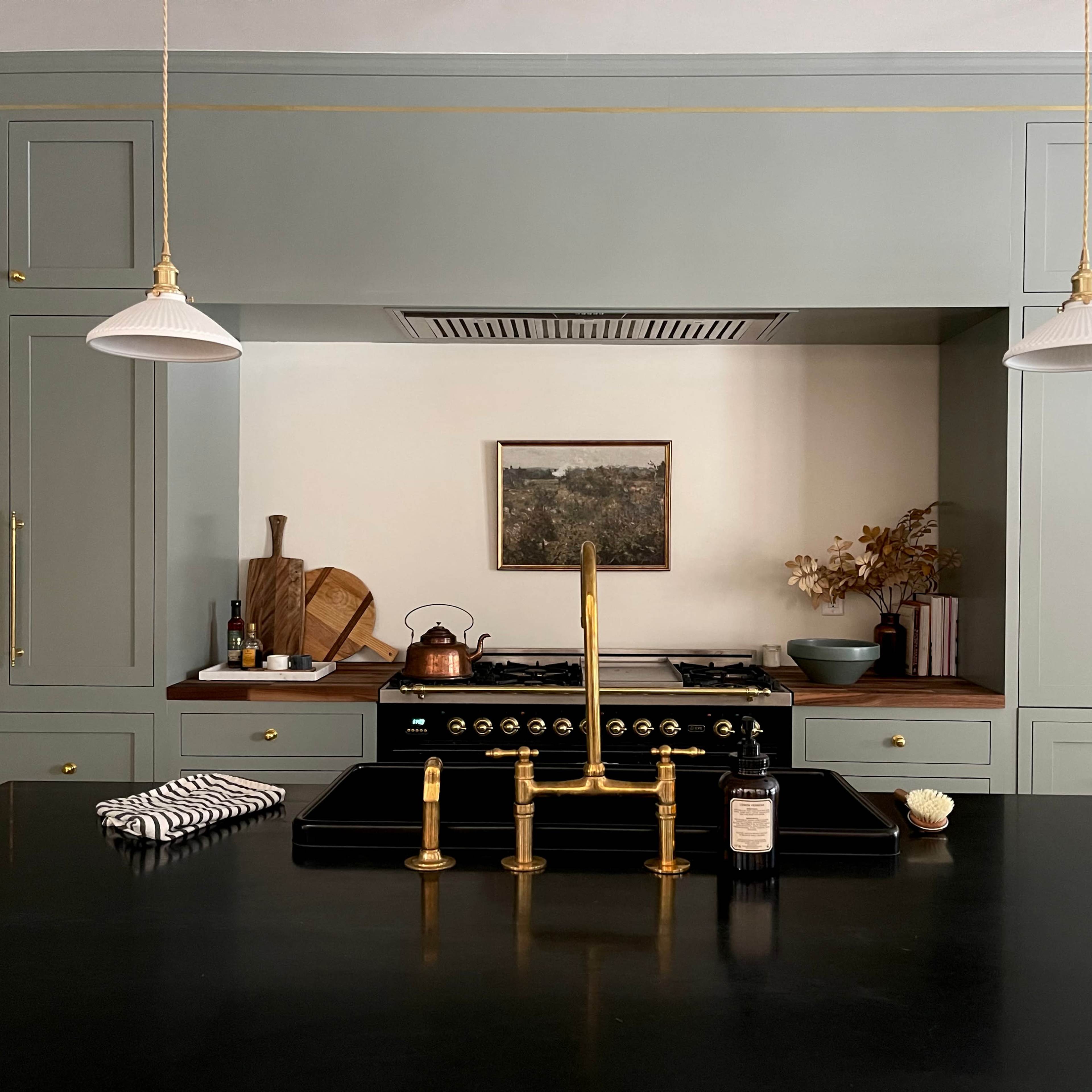 A modern kitchen features green cabinetry, a black countertop with a brass faucet, hanging light fixtures, and various kitchen utensils.